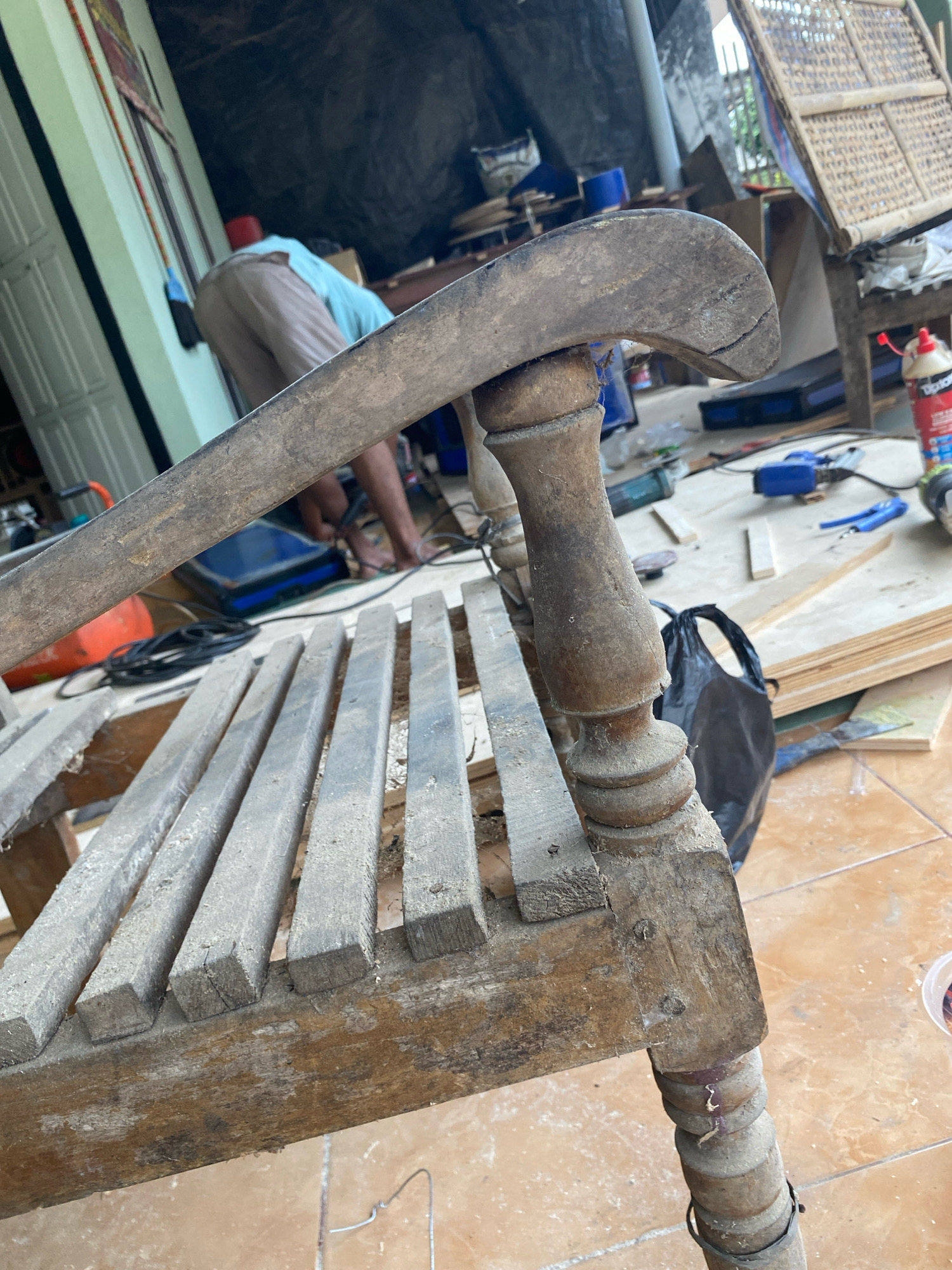 This is a close-up, eye-level shot of a weathered wooden chair undergoing repair in an outdoor workshop setting in Mojo Pahit, Indonesia. The chair, with its slatted seat and intricately carved legs and armrest, is dusty and shows signs of age and wear, including cracks in the wood. In the background, a person wearing a light blue shirt and light-colored pants is bent over, seemingly engaged in work. Various tools and materials are scattered around, including what appears to be a glue bottle, power tools, pieces of wood, and a black plastic bag, suggesting an ongoing restoration project. The ground is tiled with a terracotta-colored pattern. The overall impression is one of active, hands-on craftsmanship in a utilitarian outdoor environment.
