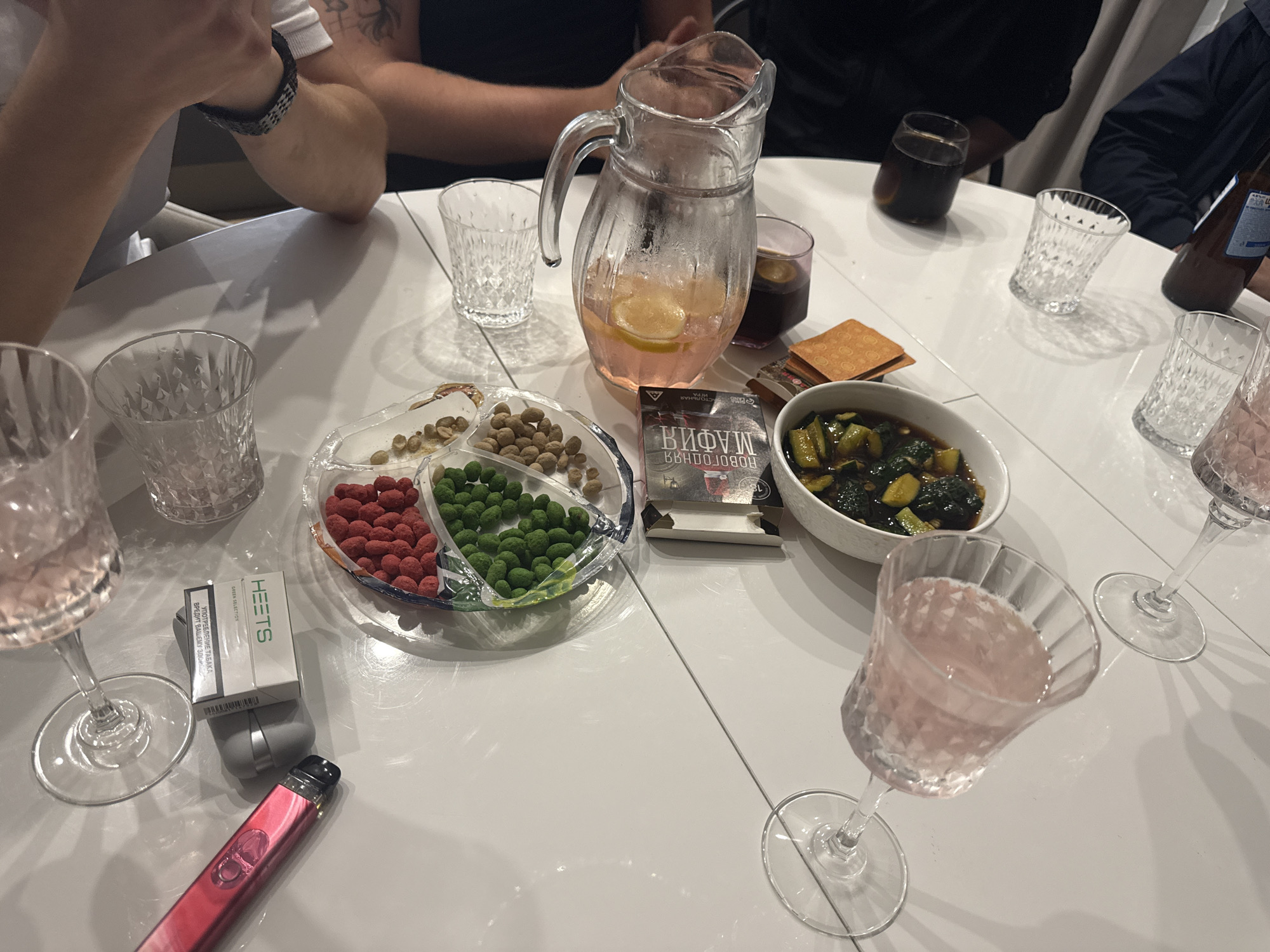 The image shows a table setting. Four people are partially visible around the table, suggesting a gathering or social event. The table is white and rectangular.
Objects on the table include:
* Several crystal glasses filled with a pink liquid.
* A pitcher containing a pink liquid with lemon slices.
* A clear glass.
* A bowl of sliced cucumbers in a dark sauce.
* A divided dish containing red, green, and brown candies/nuts.
* A pack of HEETS (tobacco sticks for IQOS).
* An IQOS device in pink.
* A box with Cyrillic text, specifically containing the word "МАФИЯ" (Mafia).
* Crackers.
* A bottle of beer.
* A dark-colored liquid in a glass.
The environment is indoors. No specific details indicate the location is in Лобаново, Russia, but the Cyrillic text on the box suggests a Russian-speaking region.
The scene suggests a casual gathering involving drinks, snacks, and possibly a game of Mafia.