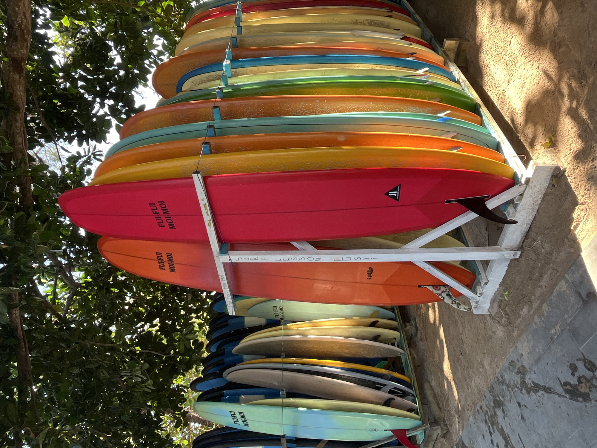 A rack of colorful surfboards stands on a sandy ground beneath the dappled shade of leafy trees. The surfboards are arranged in a row, showcasing a vibrant spectrum of colors, including red, orange, yellow, green, blue, and teal. Some boards have writing on them, such as "FUI-FUI MOI-MOI" and "SURF LESSON". The rack is a white, wooden structure. In the foreground, there is a paved area with water reflections. The scene appears to be outdoors, likely in a beachside location, possibly in Kuta Selatan, Indonesia, given the presence of surfboards. The lighting suggests it is daytime, with sunlight filtering through the trees. No people are visible in the image.