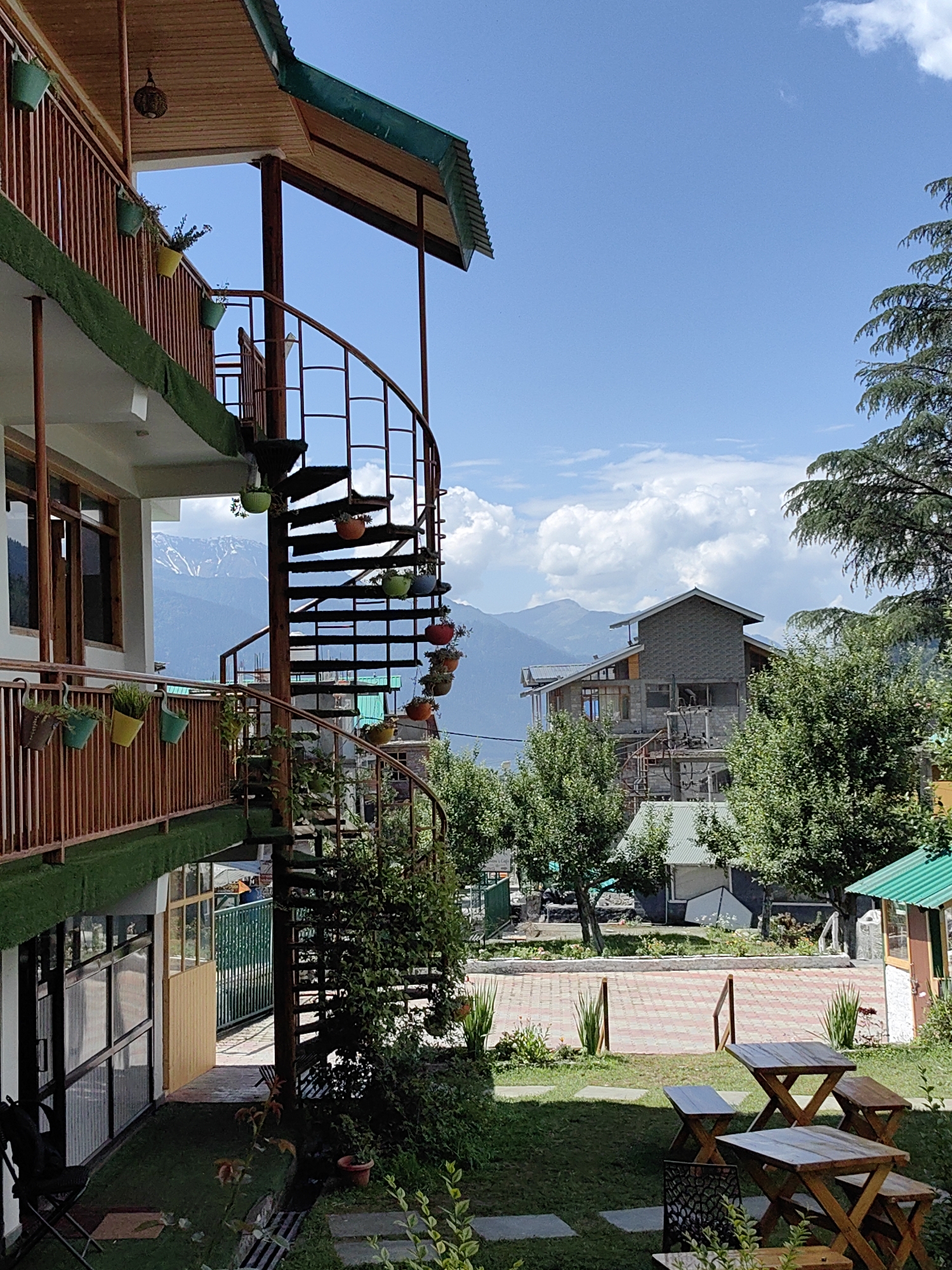 This is a daytime, eye-level shot of a hotel or guesthouse in Vashisht, India. The building's exterior features a wooden balcony with a railing adorned with several potted plants. A spiral staircase made of dark metal descends from an upper level. The background reveals a mountainous landscape under a bright blue sky with scattered clouds. In the middle ground, other buildings and lush green trees are visible. The foreground shows a grassy area with several wooden picnic tables and benches. The overall impression is of a serene, outdoor setting in a hilly region.