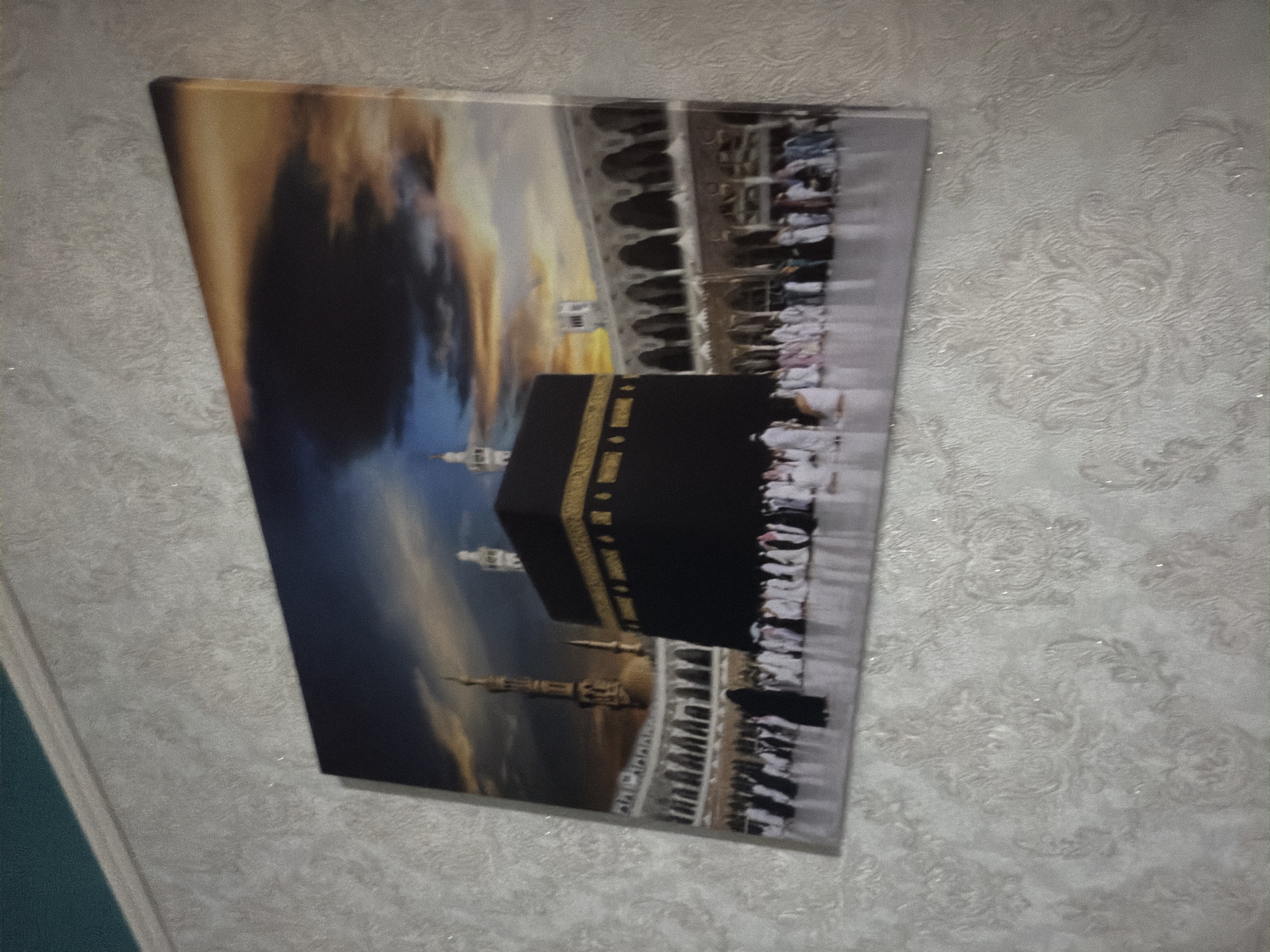 A canvas print depicts the Kaaba in Mecca, Saudi Arabia. The image shows the black cube-shaped structure with a golden trim, surrounded by white marble buildings with arched walkways. A crowd of people, many wearing white ihrams, are gathered around the Kaaba. The sky above is a dramatic mix of dark clouds, blue sky, and orange and yellow hues, suggesting either sunrise or sunset. The print is hung on a textured, light-colored wall with a subtle floral pattern. The wall appears to be in a residential setting, possibly a living room, in an unknown location in Russia.