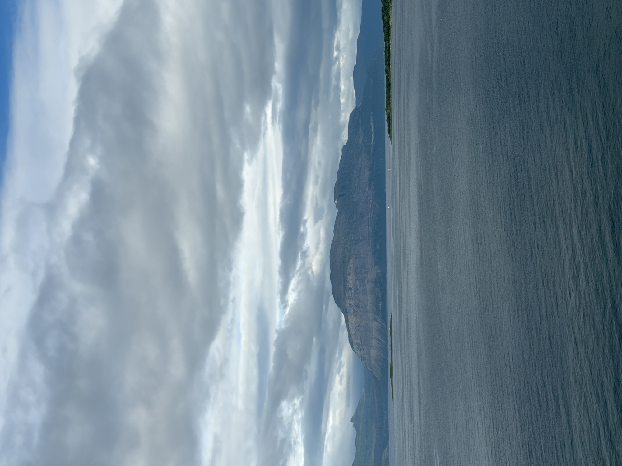 The image depicts a serene landscape under a cloudy sky. The foreground is dominated by a vast, calm body of dark blue water, with subtle ripples indicating a gentle current. In the middle ground, a narrow strip of green land forms an island, topped with a small white structure. Beyond the island, a larger landmass rises, characterized by rugged, layered mountains with a dark, rocky texture. The mountains are partially obscured by clouds, with hints of snow on their peaks. The sky is a dramatic canvas of grey and white clouds, with patches of blue peeking through. The overall scene suggests a remote, natural environment, likely a fjord or coastal area. While the image does not directly show the city of Harstad, Norway, such a landscape is characteristic of the region, where fjords, mountains, and islands are common features, often with weather patterns similar to those depicted.