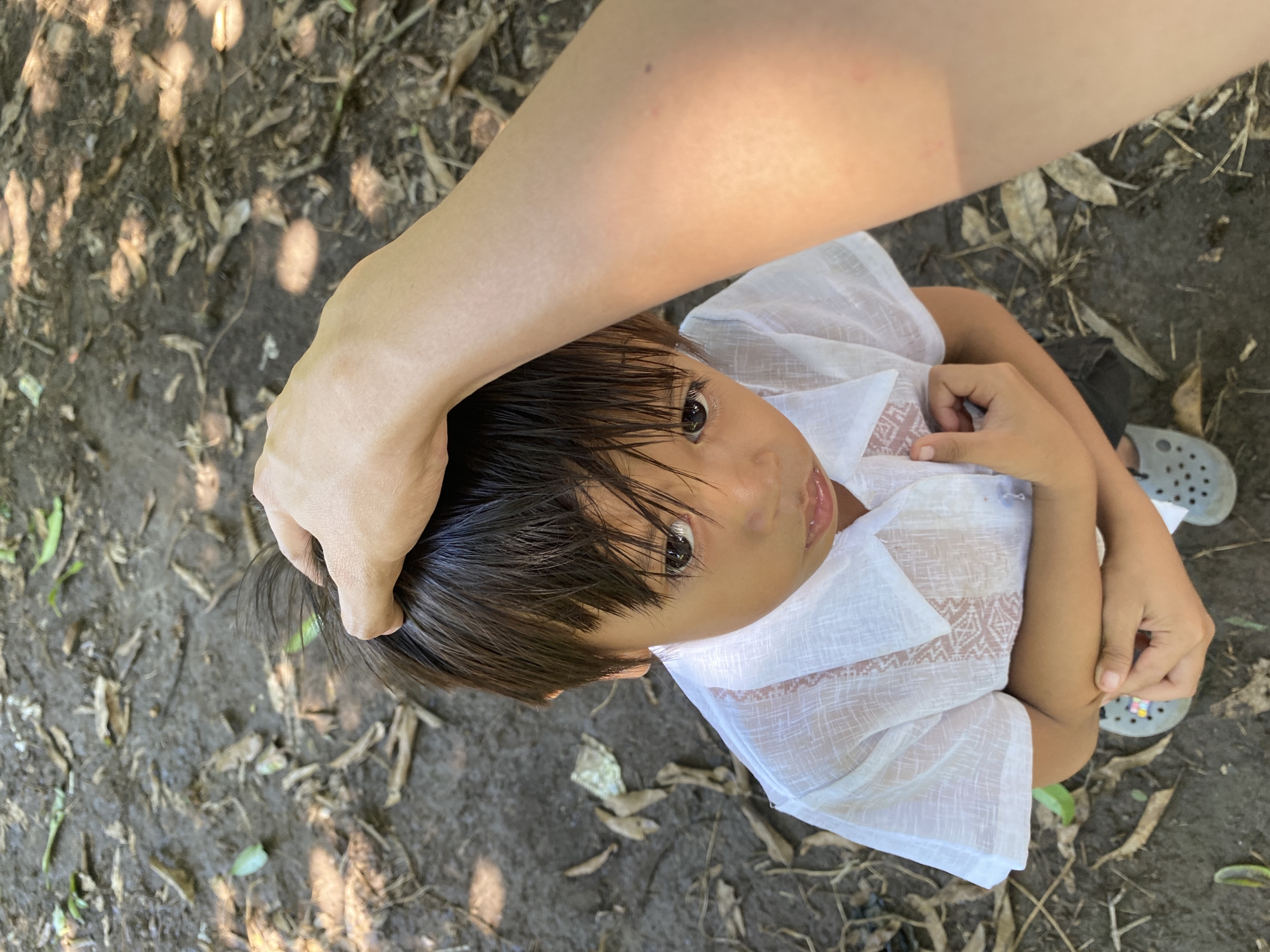 A child, appearing to be of Southeast Asian descent, is captured from a high-angle perspective outdoors. The child is wearing a white collared shirt and dark shorts, with gray footwear visible. Their hands are clasped together in front of them, and they are looking upwards towards the camera with a somber expression. A hand, presumably belonging to the photographer, is gently resting on the child's head. The ground beneath them is a mixture of soil and fallen leaves, suggesting a natural or park-like setting. The dappled sunlight filtering through overhead foliage indicates this is likely a shaded area in a tropical or subtropical climate. The presence of foliage and the type of ground cover are consistent with outdoor environments found in Davao City, Philippines, such as local parks, schoolyards, or residential areas with natural landscaping.