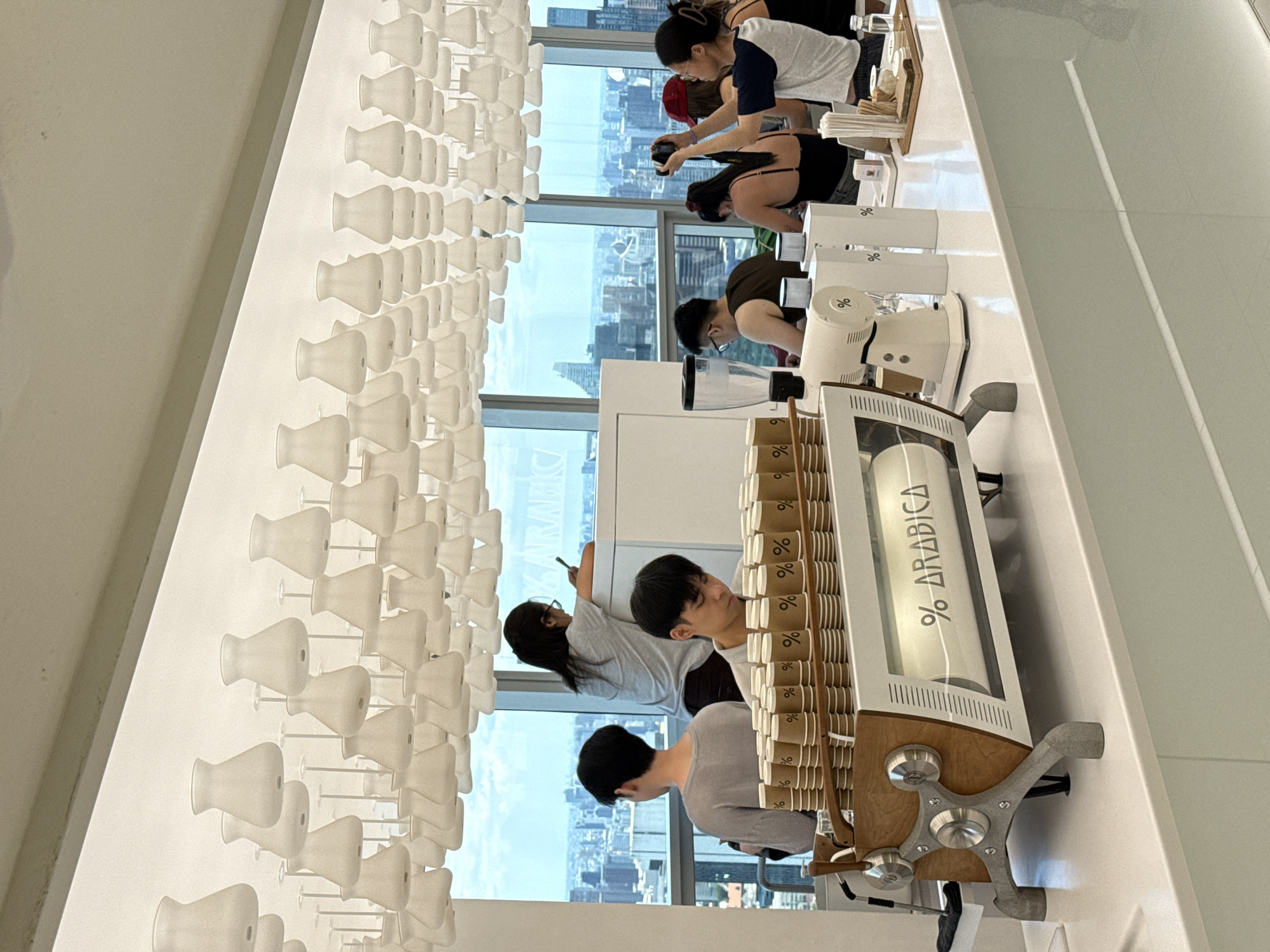 The interior of a cafe features a long white countertop with customers and employees. A row of patrons faces windows overlooking a city skyline, likely Bangkok, Thailand, given architectural clues. Employees stand behind a coffee machine labeled “% Arabica” and stacks of branded coffee cups. A white wall is decorated with a grid of small, white, goblet-shaped fixtures. Individuals are standing, interacting, and photographing the scene with their phones.