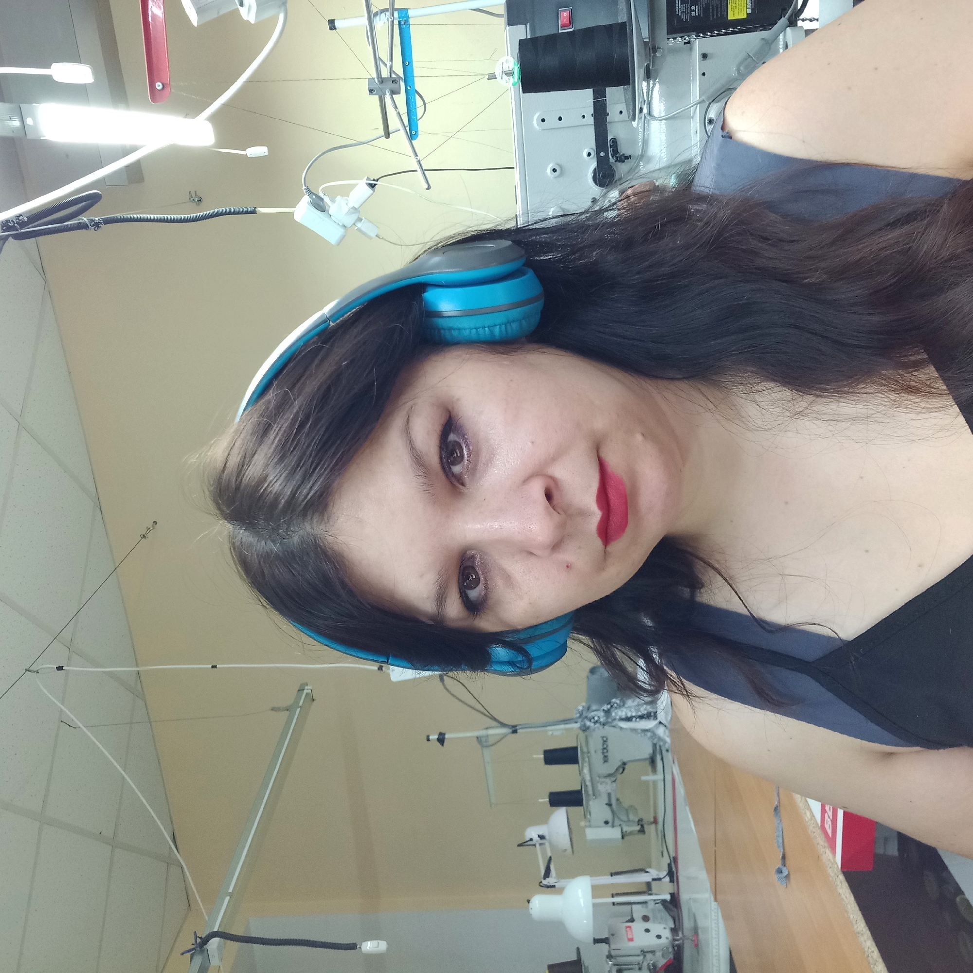 The image is a selfie taken indoors, likely in a workshop or factory setting, given the presence of sewing machines and industrial lighting. A woman with dark hair and blue headphones is in the foreground, looking at the camera. She is wearing a dark top. In the background, several sewing machines are visible, along with spools of thread and various industrial equipment. The lighting appears to be a mix of overhead fluorescent lights and task lighting. There is no indication that this image was taken in Kropyvnytskyi, Ukraine.