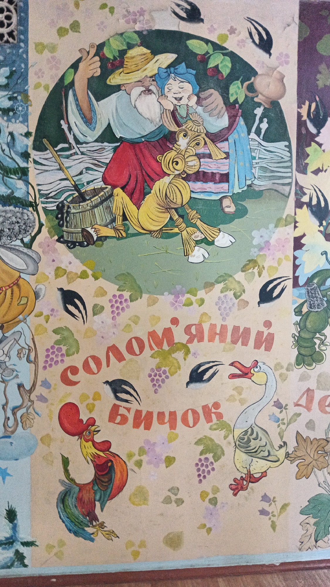 The image is a close-up of a colorful mural or artwork, likely on a wall or large surface. The style is illustrative and folk-art inspired, featuring characters and decorative elements.
In the upper portion of the image, a man with a white beard and a straw hat is embracing a woman with a blue headscarf and closed eyes. They appear to be a couple, possibly a farmer and his wife, depicted in traditional attire. Between them and slightly below, a yellow, straw-like figure resembling a scarecrow or a stylized character is lying down. Behind the couple, there are green leaves, red berries, and a wooden fence. In the background, a wooden bucket with a handle and a ladle are visible. Several small black birds are depicted flying in the upper section.
The lower portion of the image is dominated by text in large, reddish-orange Cyrillic letters. The visible text reads "СОЛОМ'ЯНИЙ БИЧОК", which translates to "Straw Bull". Around the text are various decorative elements including clusters of purple grapes, small pink and purple flowers, green leaves, and more small black birds in flight.
To the left of the text, a brightly colored rooster with a red comb and a vibrantly colored tail is standing. To the right of the text, a white goose with yellow feet and beak is depicted. Further to the right, partially visible, is a green lobster or crayfish figure.
The background of the entire artwork is a light, speckled beige color. The overall impression is a scene from a fairy tale or folk story, executed in a vibrant and charming artistic style. The location of this artwork is on the exterior wall of a building at 35 Prospekt Lenina (now Prospekt Myru) in Kryvyi Rih, Ukraine, specifically on the wall of the Kryvyi Rih Academic Ukrainian Music and Drama Theater.