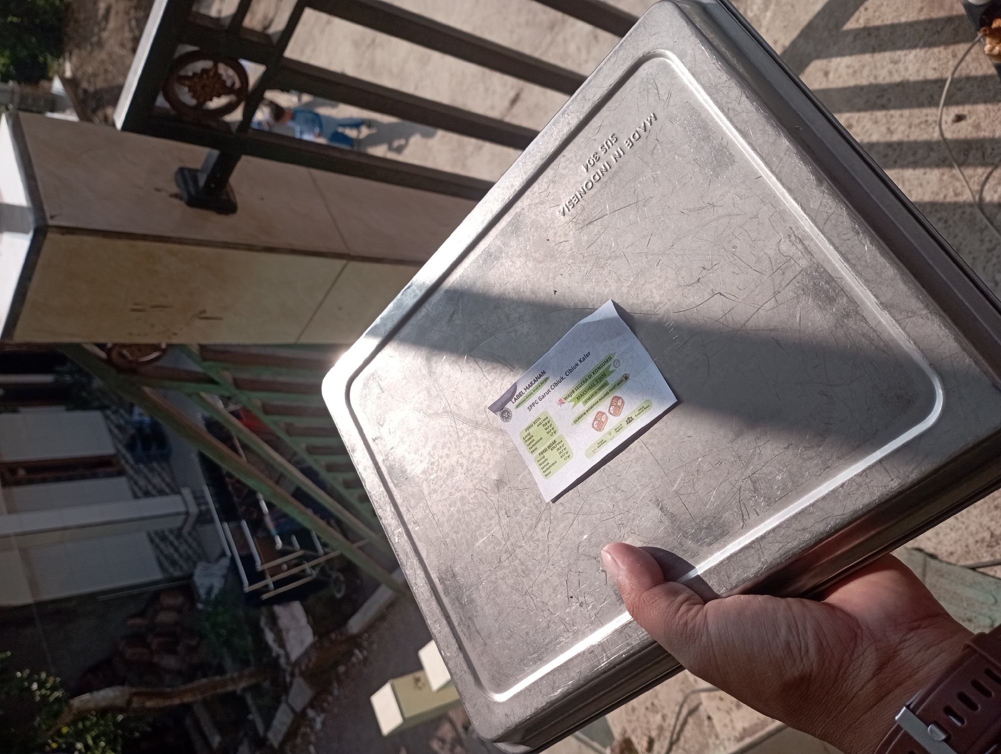 A person's hand holds a rectangular metal baking pan. The pan has visible scratches and wear marks. A small white label with text and images is attached to the pan's surface. The label contains information about food ingredients and nutritional content, including text in Indonesian. The background shows a building with a tiled facade, a dark green metal fence, and a paved area. The lighting suggests it is daytime, with strong sunlight casting shadows. The location is identified as Garut, Indonesia, by the text on the label which reads "SPPU: Garut Cihui, Cibiru, Kafer".