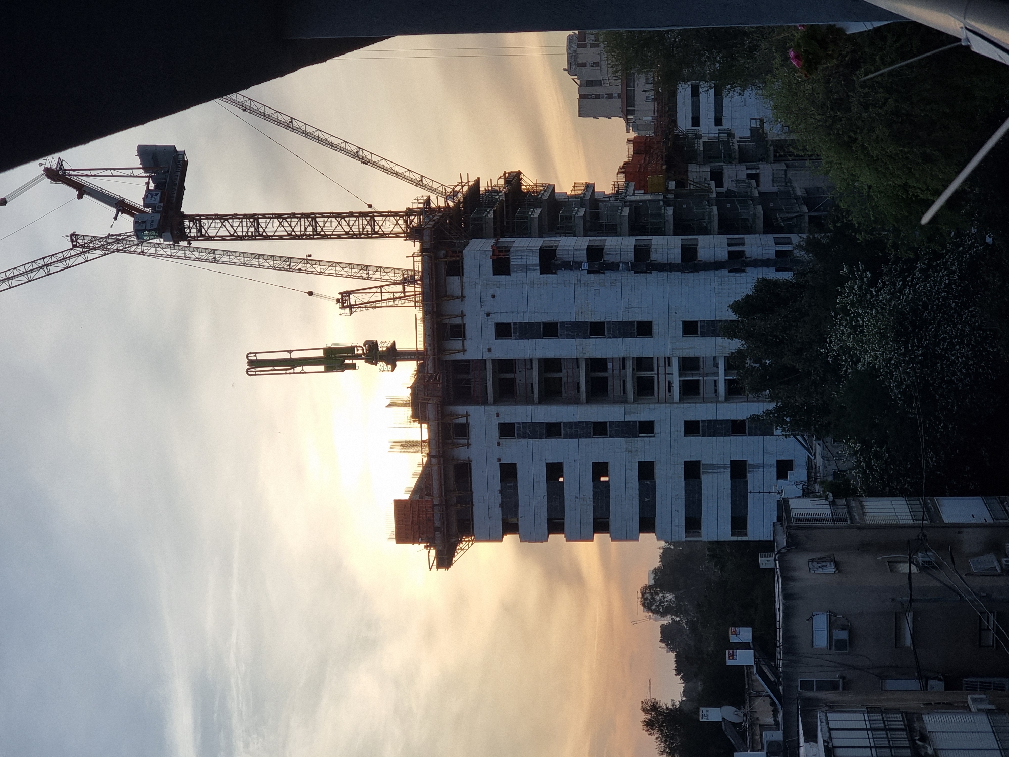 The image shows a construction site in Ramat Gan, Israel, with a high-rise building under construction. Several cranes surround the building. The sky is a mix of orange and yellow, suggesting either sunrise or sunset. In the foreground, a building with multiple windows and air conditioning units is visible, potentially residential. Trees and some greenery are interspersed between the buildings. The scene appears to be viewed from a high vantage point, possibly from another building.