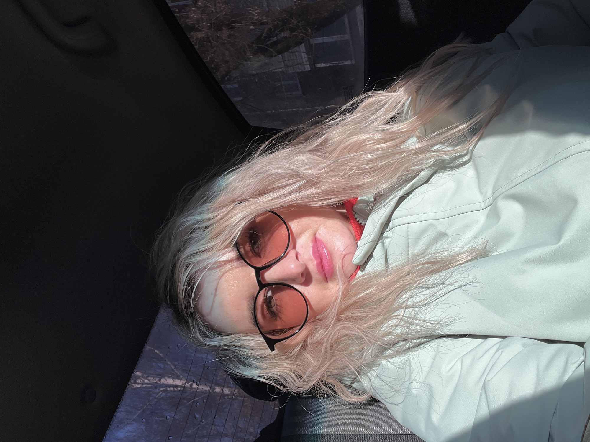 A woman with long, wavy, light blonde hair, which appears to have a subtle pink tint, is featured in a close-up, angled portrait taken inside a vehicle. She wears large, round sunglasses with black frames and orange-tinted lenses, through which her eyes are faintly visible. Her lips are adorned with pink, glossy lipstick or gloss, and she has a calm, slightly smiling expression.
She is dressed in a light pastel green or light grey zippered jacket, possibly made of a synthetic, weatherproof material, with a hint of a red garment visible underneath, perhaps a hood or collar. The bright, direct sunlight streams from the left, casting prominent highlights on her hair and jacket, and creating shadows on her face and the car's interior.
The setting is clearly inside a car, with reflections in the upper car window or sunroof revealing bare tree branches and distant structures, suggesting an outdoor, possibly urban or suburban environment during the daytime. Dark car interior elements, including a textured seat or dashboard, are visible at the bottom right. The overall activity appears to be a relaxed selfie or portrait taken while seated in the car. There is no visible text in the image.