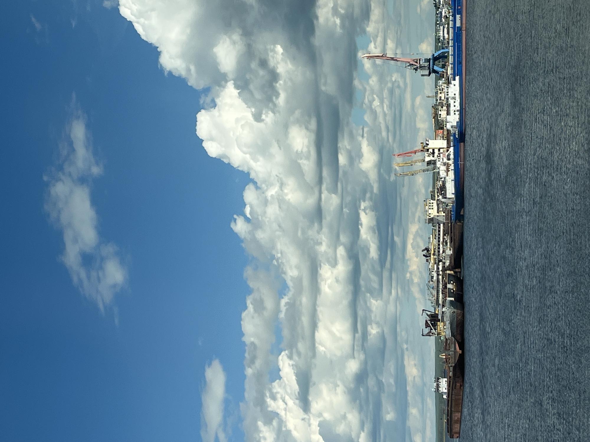 The image depicts a view of the Kama River in Chaikovsky, Russia, showing a river port facility under a cloudy sky. The left side of the frame features blue sky and large cumulus clouds. On the right, a body of water occupies a significant portion, likely the Kama River. Along the riverbank are various ships and port infrastructure, including cranes and buildings. The cranes are of different sizes and colors (red, yellow, blue). The ships appear to be docked, with multiple structures and equipment visible. The horizon line is not distinctly visible, but seems to blend with the clouds and the urban landscape. The overall scene suggests industrial activity at a river port.