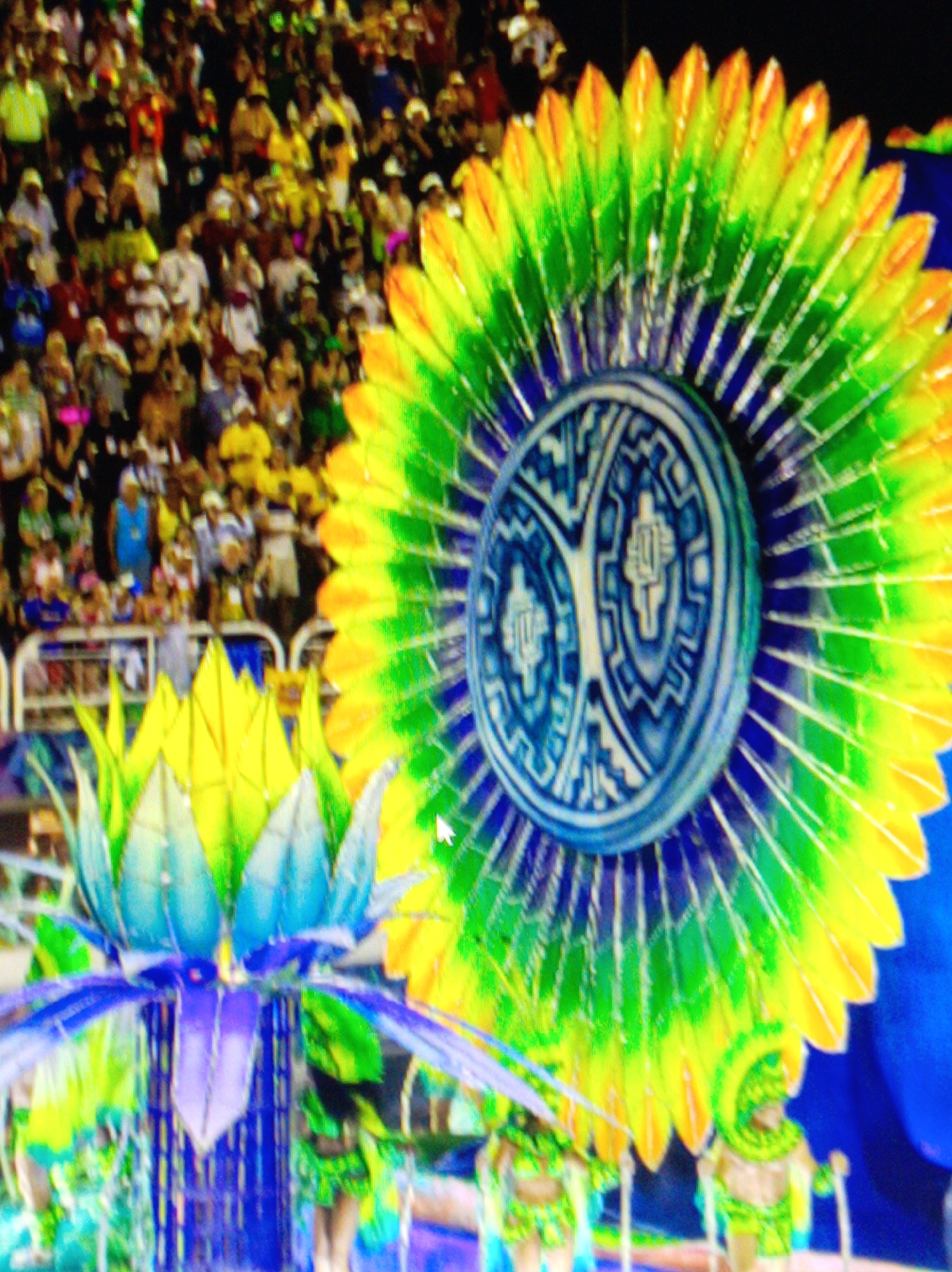 In a stadium, a large crowd of people are watching a parade. The parade features elaborate costumes and floats. One of the main subjects is a large, elaborate headdress or fan, decorated with vibrant green, yellow, and blue feathers. In front of this headdress is a smaller, blue and purple flower-like structure. In the background, people are visible in the stands and some performers in costumes can be seen on the parade route, holding staffs. The overall scene suggests a festive event, likely a carnival or parade. The stadium setting implies an organized public gathering.