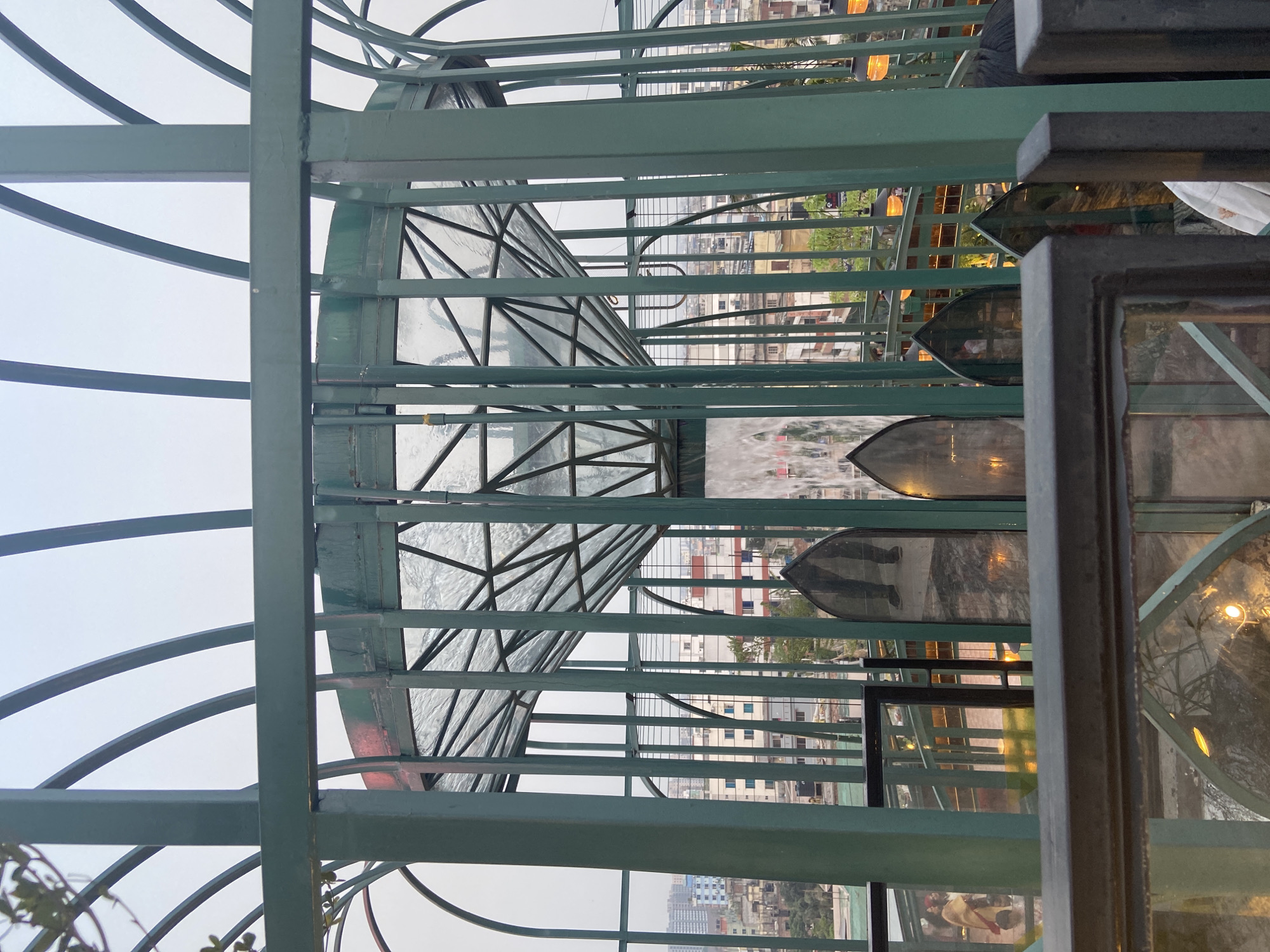 The image shows a green metal structure with glass panels, likely part of a rooftop or balcony enclosure, in Dhaka, Bangladesh. Through the metal bars and glass, a cityscape of Dhaka is visible with buildings of varying heights and density. Inside the structure, there are reflective surfaces, possibly mirrors or glass panels, that distort the urban landscape. The overall atmosphere is slightly muted due to the overcast sky.
