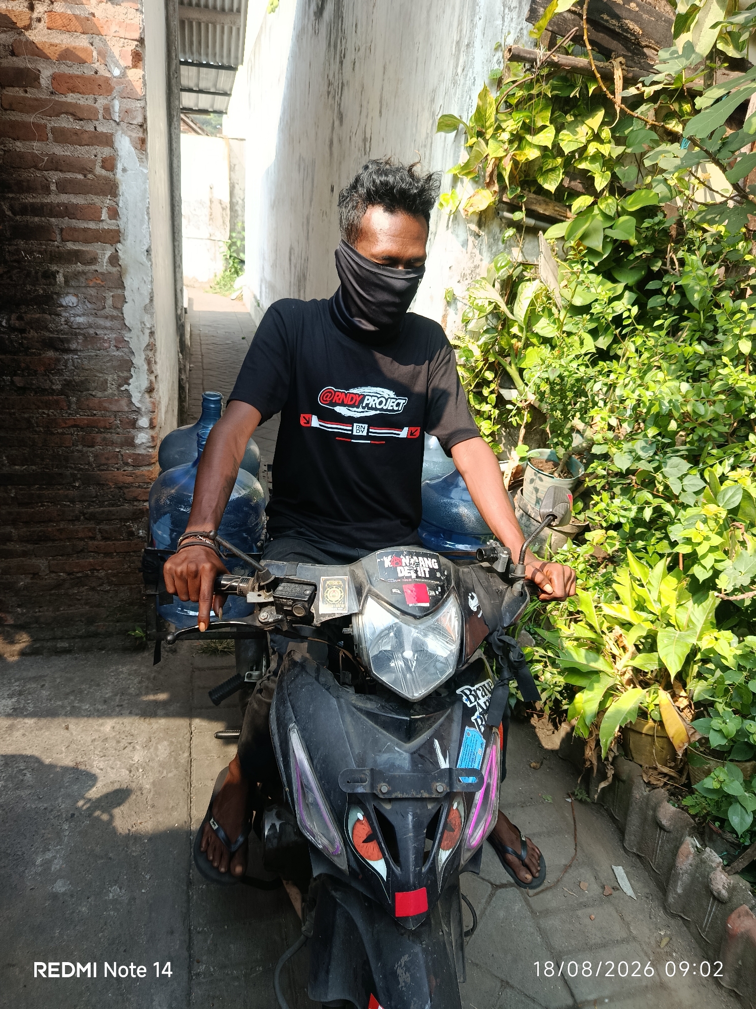 A man wearing a black face mask and a black t-shirt with a white and red logo is sitting on a black motorcycle in a narrow alley. He is holding the motorcycle's handlebars with his right hand. On the back of the motorcycle, there are two large blue water jugs. The alley has brick walls on the left and a white wall with plants on the right. The ground is paved with interlocking bricks. The lighting suggests it is daytime. A timestamp in the lower right corner reads "18/08/2026 09:02". A watermark in the lower left corner reads "REDMI Note 14".
