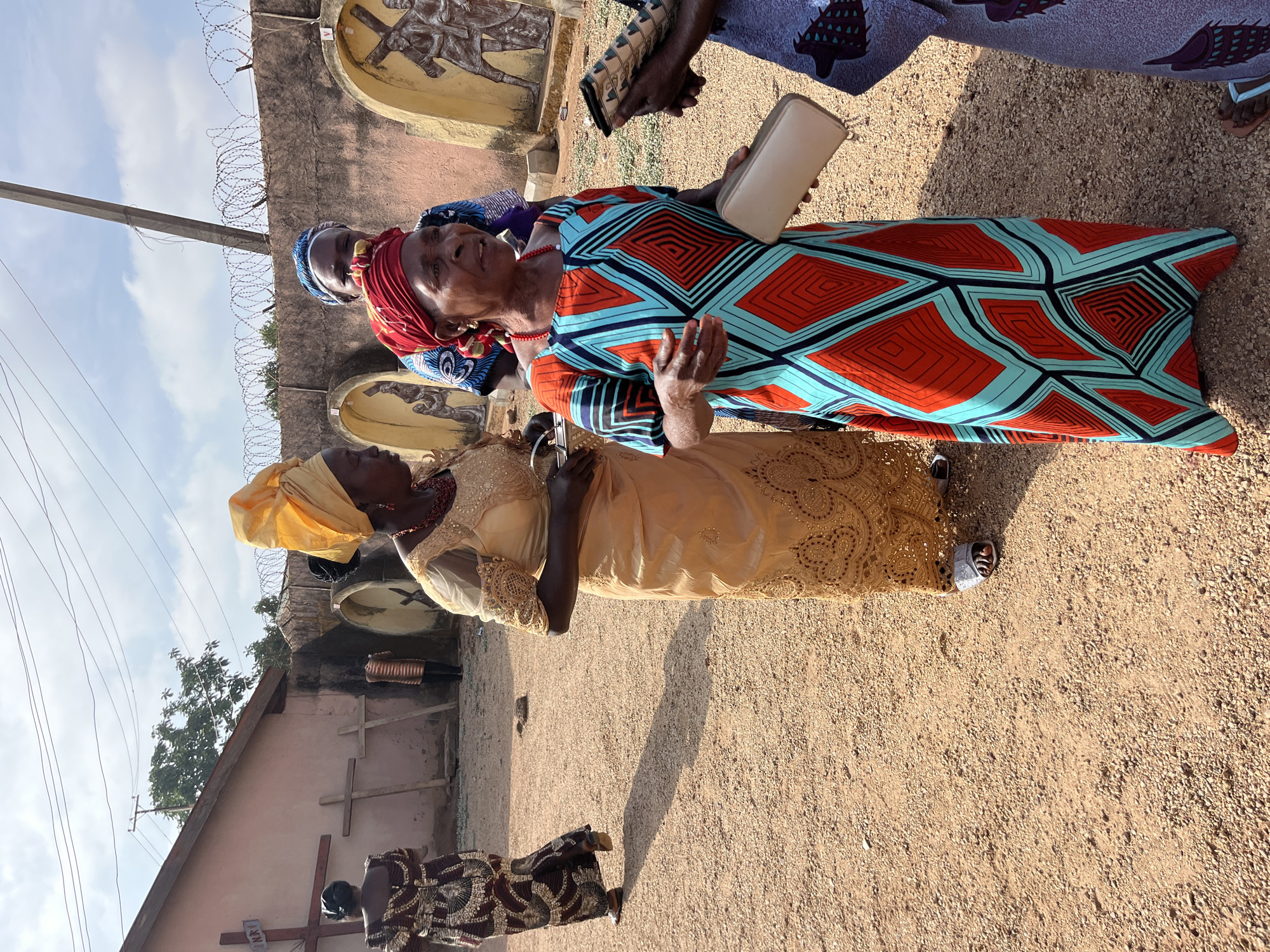 Three elderly African women stand on a dirt surface with gravel. The woman closest to the viewer wears a yellow headscarf and a gold-colored lace dress. The woman in the middle wears a red headscarf, a dress with an orange and turquoise geometric pattern, and holds a white purse or book. A third woman stands behind her wearing a blue patterned headscarf. Behind the women is a light-colored wall topped with barbed wire and featuring decorative carvings of figures on crosses. A building is visible in the background to the left with a cross on its exterior. The photograph appears to have been taken outdoors in Jos, Nigeria.