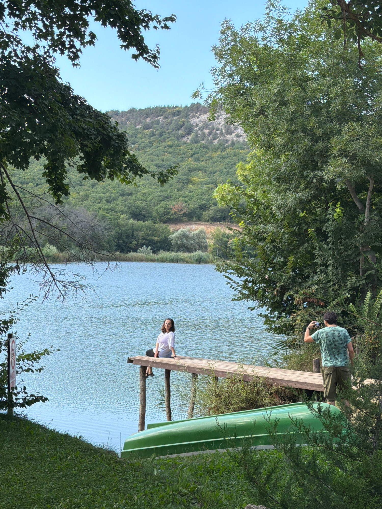 The image depicts a serene natural scene by a body of water, likely a lake or calm river, with a wooded hillside in the background. In the foreground, a green boat rests on grassy terrain. A wooden pier extends into the water, where a young woman is seated, dressed in a white shirt and dark shorts. To the right of the pier, a man wearing a green camouflage t-shirt and khaki shorts is standing, holding a camera and appearing to take a photograph. The environment is lush with trees and vegetation, and the sky is clear and blue. The location is identified as being in Balaklavsky District, Ukraine.
