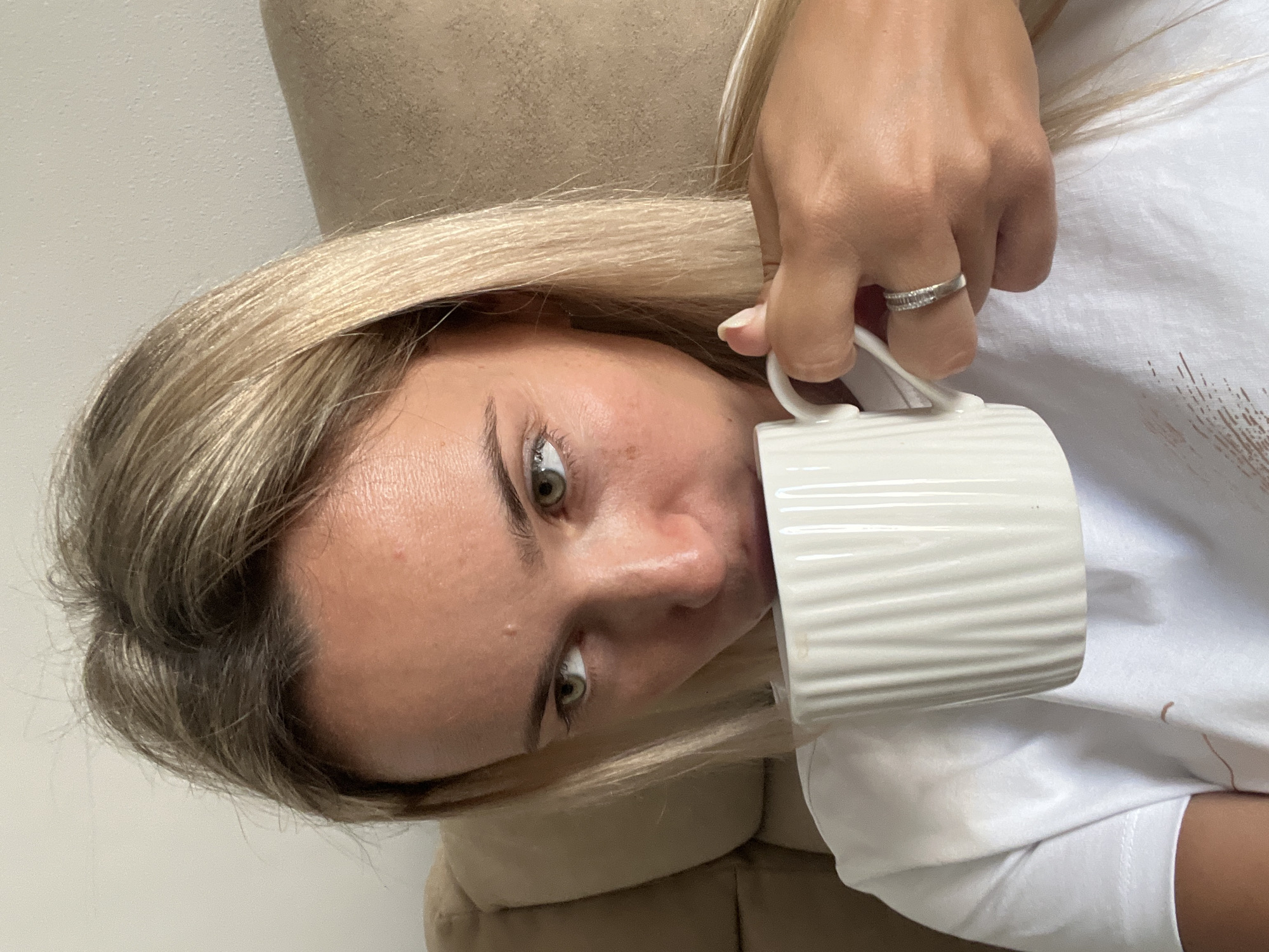 A young woman with blonde hair and green eyes is holding a white mug and drinking from it. She is wearing a white t-shirt and a ring on her finger. She is sitting on a couch, which appears to be in a home or hotel room. The background is neutral, with a hint of a wall and the back of the couch. The lighting is natural and appears to be from a window. The image does not contain any information or visual cues that would indicate the location is Sharm El Sheikh, Egypt.