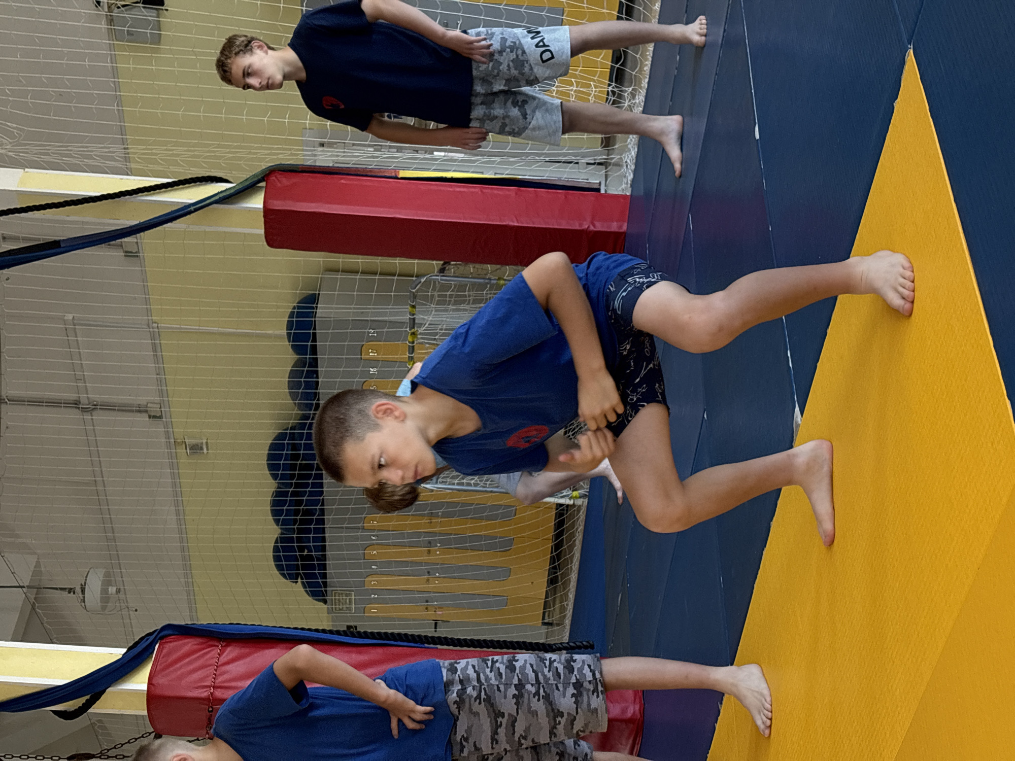 The image depicts a wrestling practice session with three young male athletes in a gymnasium. The setting includes wrestling mats, a red wrestling mat, and a protective net. The athletes are dressed in wrestling attire, with two of them wearing dark blue t-shirts and camouflage shorts, while the third is in a blue t-shirt and patterned shorts.
One athlete is standing and looking down, seemingly observing the action. Another is in a crouched position, possibly demonstrating a wrestling move. A third athlete is visible at the bottom of the frame, also in athletic wear. The flooring is a combination of blue and yellow wrestling mats, suggesting a dedicated training area.
There are some visible markings on the wall, including what appear to be numbered ranks or progress markers, with numbers ranging from 1 to 7. The lighting suggests an indoor environment, likely during daytime. The overall atmosphere appears to be one of focused training and physical activity. The cropped images highlight details of the athletes' attire and the mat textures. No specific emotions are overtly displayed, but the context implies concentration and effort. No visible text is present beyond the numbered markings on the wall. The location is Chaikovsky, Russia.