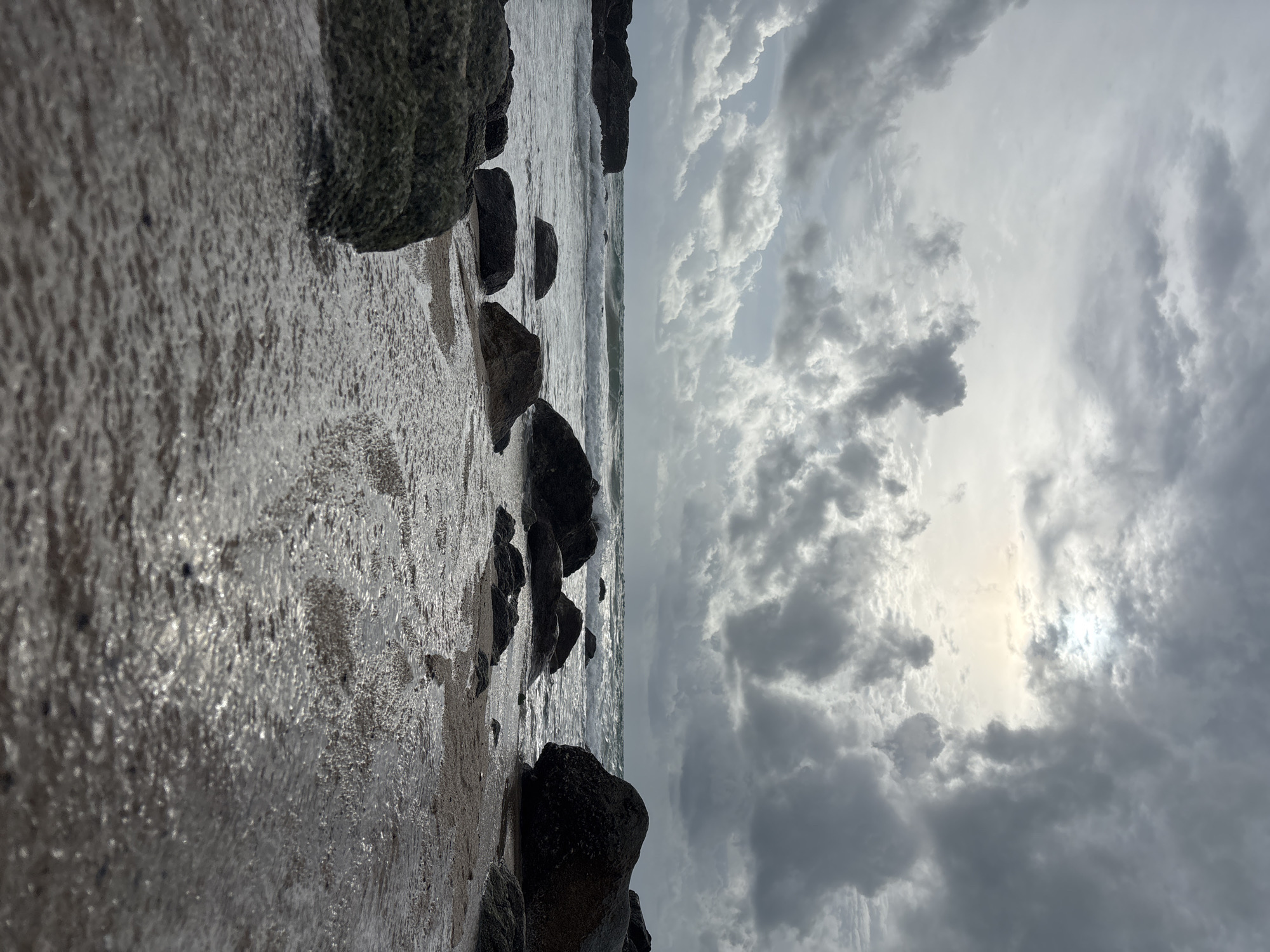 The image shows a rocky beach at Choeng Thale, Thailand, under an overcast sky. The foreground is dominated by large, dark rocks along the shoreline, partially submerged in water. The water itself is churning slightly, with small waves visible. The sky is filled with dense, gray clouds, obscuring the sun. The scene appears devoid of people.