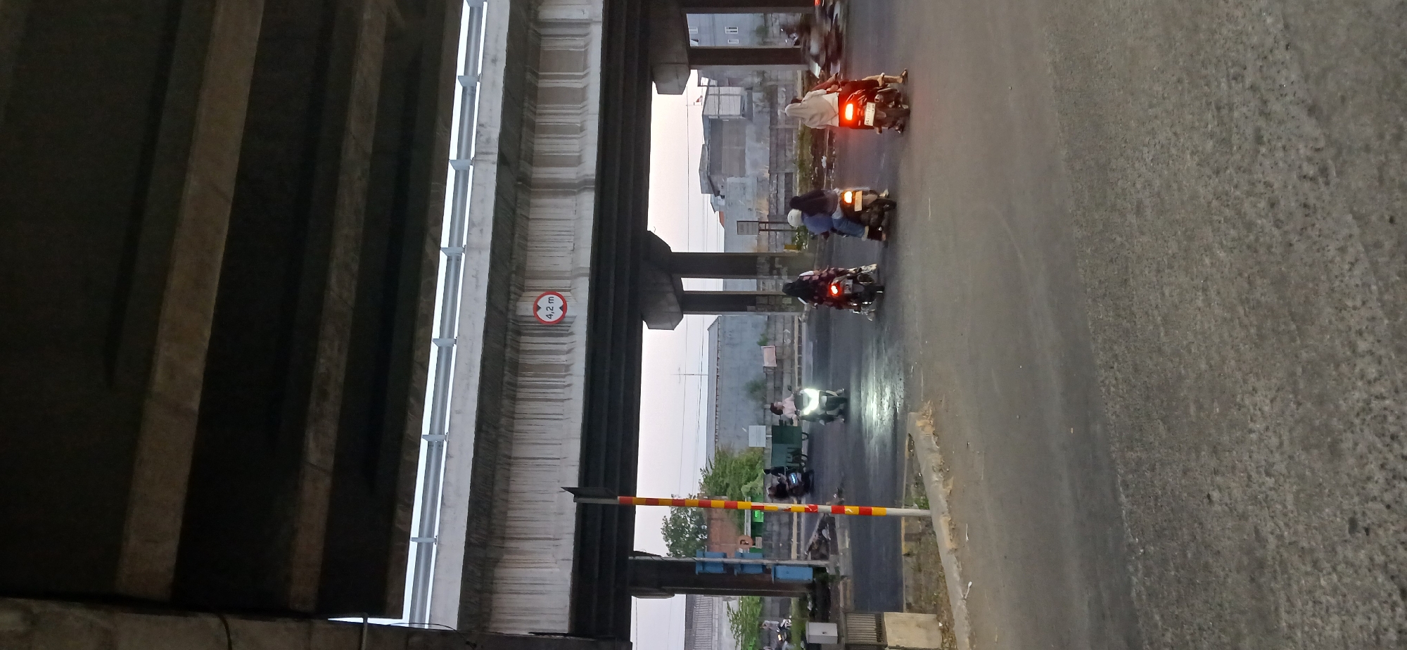 In Wadungasri, Indonesia, beneath a concrete overpass, several individuals ride motorcycles on a road at dusk. A sign on the overpass indicates a clearance height of 4.2 meters. Four motorcycles are visible, carrying a total of six people. One motorcycle in the foreground is ridden by a person wearing a white headscarf and a white shirt. Another motorcycle behind it has two occupants, one wearing a blue jacket. Further back, a motorcycle with two riders is seen, both wearing dark clothing and headscarves. The motorcycle furthest to the left is ridden by a single person. The road surface is asphalt, and there are buildings and vegetation in the background. The lighting suggests early evening or late afternoon.