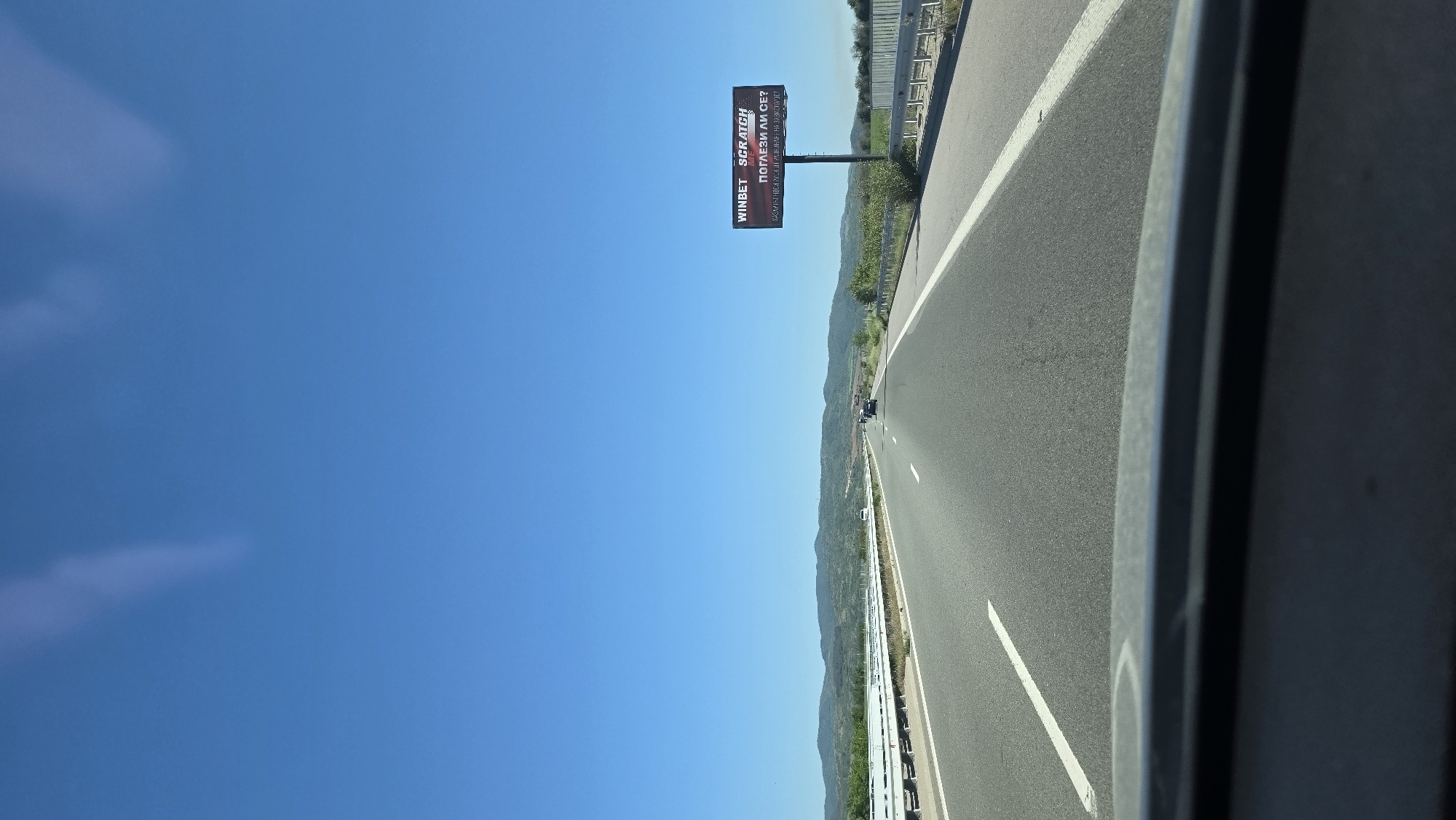 The image depicts a highway in Mursalevo, Bulgaria, under a clear blue sky. A roadside billboard displaying the "WINBET SCRATCH" advertisement stands along the side of the road. The asphalt highway has clearly marked white lines. The scene is captured from inside a vehicle with a view out onto the road and countryside.