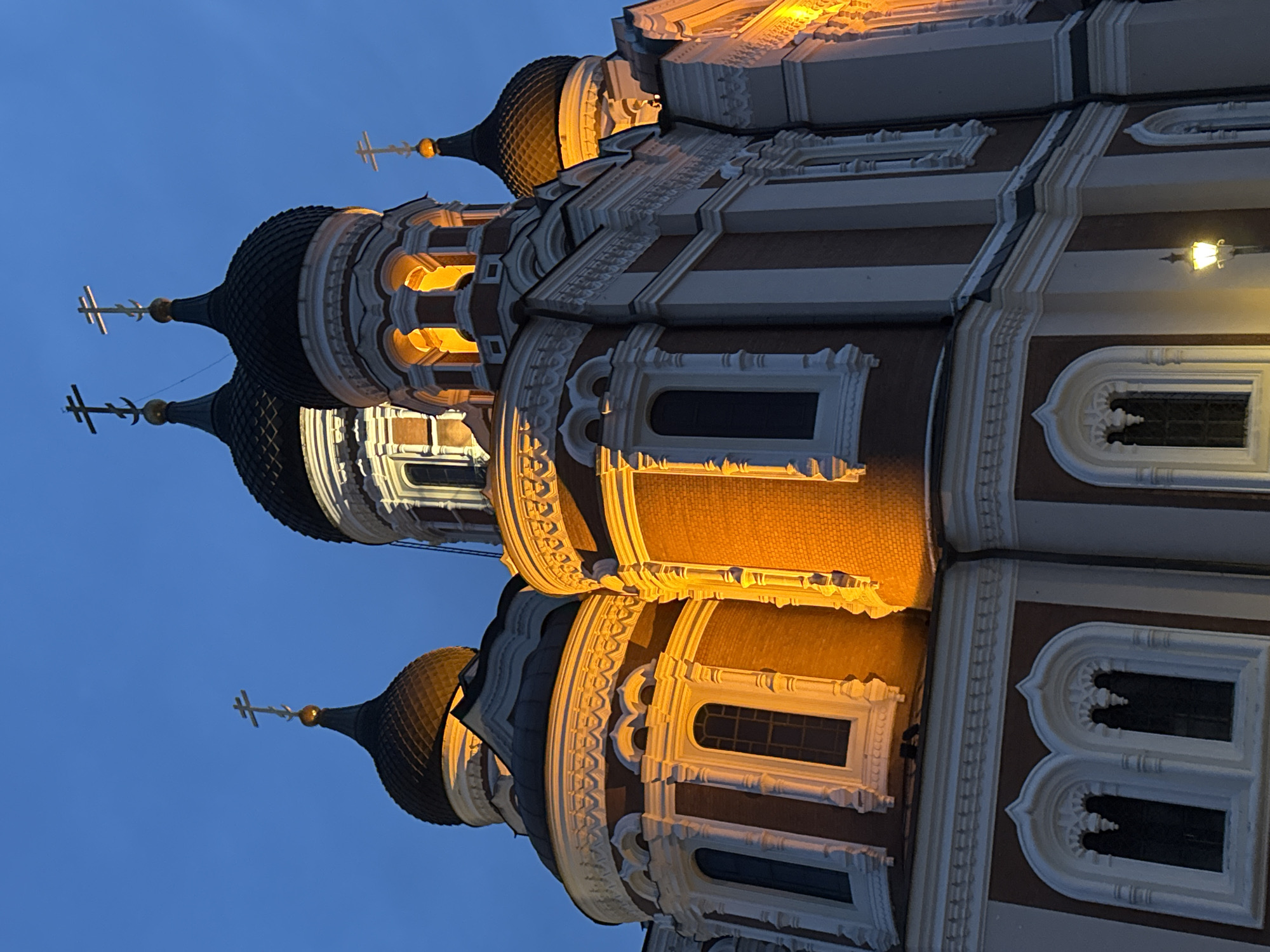 The image shows a close-up, low-angle shot of the Alexander Nevsky Cathedral in Tallinn, Estonia, at dusk. The cathedral's onion domes, topped with Orthodox crosses, are prominent against a darkening blue sky. The brick facade is illuminated by warm, artificial lighting, highlighting the ornate architectural details, including arched windows and decorative cornices. The lower portion of the building features a row of intricately carved windows. A single ornate street lamp with a glowing bulb is visible in the lower right corner. The scene is devoid of people, and no actions or interactions are depicted. The focus is on the monumental architecture of the cathedral bathed in evening light.