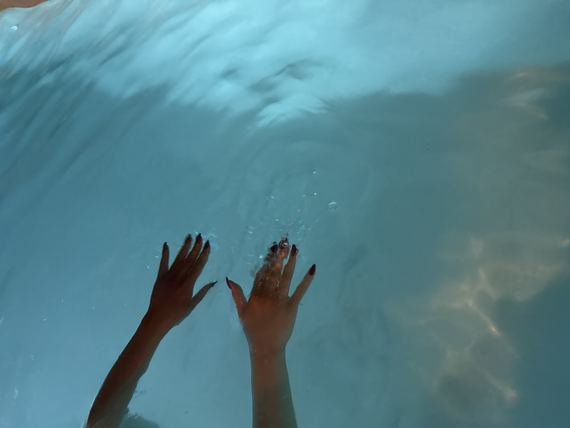 The image presents an overhead view of two human hands submerged in clear, light blue water. Both hands, positioned in the lower-central part of the frame, display long, pointed fingernails painted in a dark color, possibly red or maroon. The left hand is positioned slightly lower and to the left, while the right hand appears to be moving, causing small bubbles and ripples on the water's surface near the fingertips.
The water itself is a bright, translucent turquoise hue, reflecting light patterns that create shimmering effects and varying shades across its surface and beneath. These light patterns suggest an overhead illumination source, possibly natural light filtering down. The overall scene conveys a sense of calm or relaxation, indicative of an activity like swimming or bathing. No other subjects, objects, or text are visible in the frame, and there are no specific cues within the image to indicate the time of day or the given location of Yerevan, Armenia.