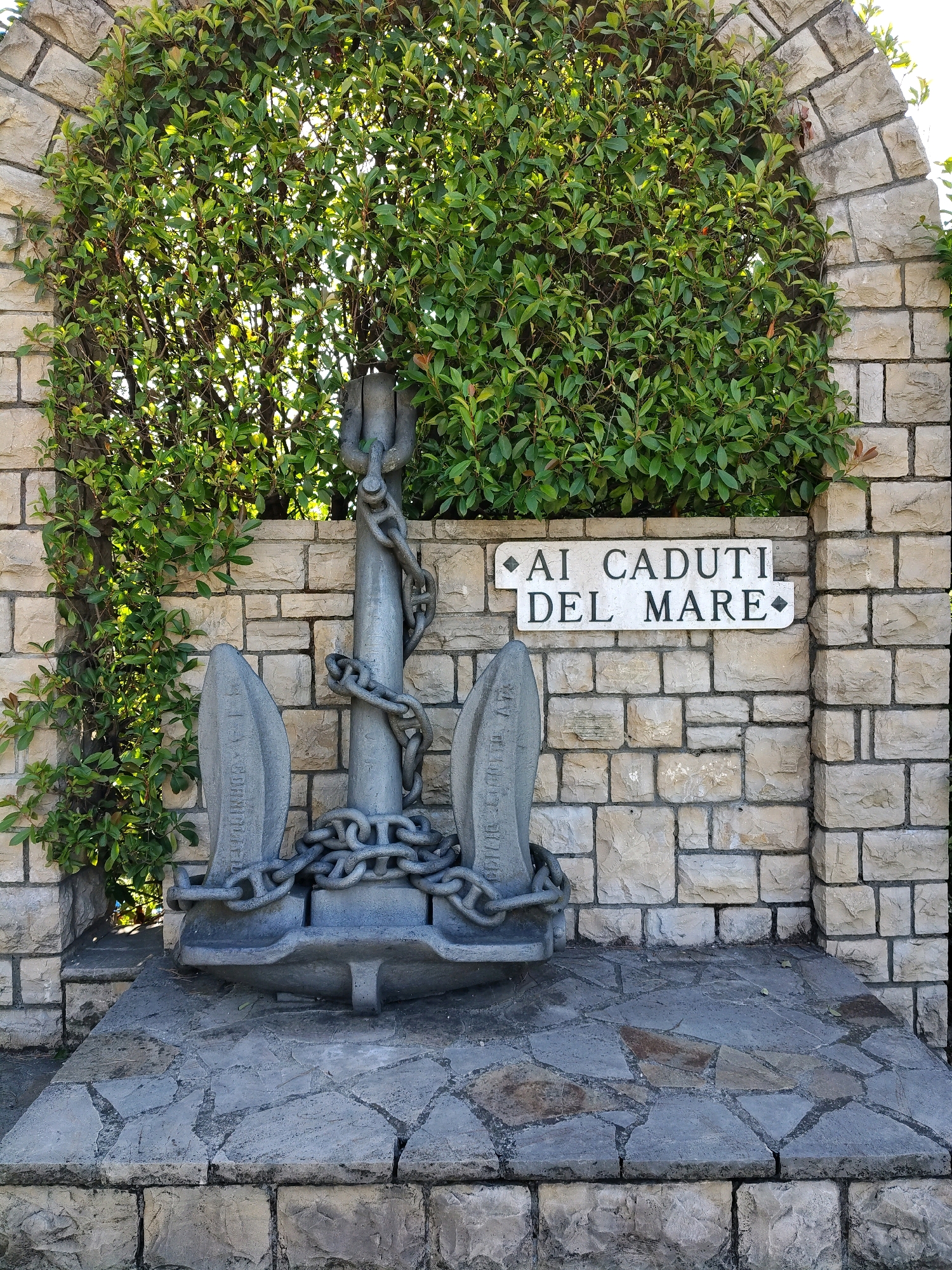 An anchor monument is centrally located in front of a stone wall covered by a dense green hedge. The anchor, made of dark gray metal, is secured by thick chains. A sign with the inscription "AI CADUTI DEL MARE" is mounted on the stone wall to the right of the anchor. The ground in front of the monument is paved with irregular gray stones. The scene is outdoors, under natural light.