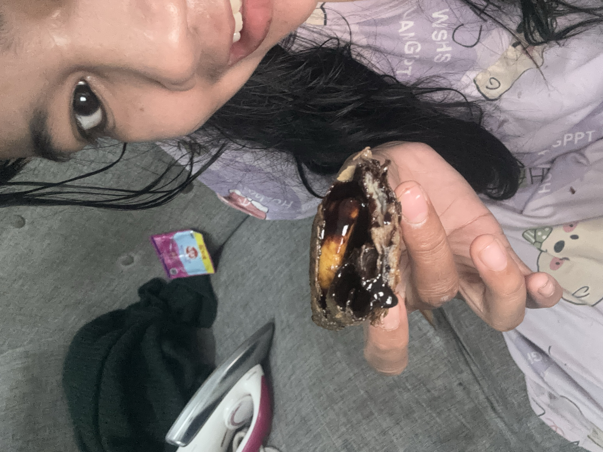 A young woman of Indonesian descent, likely in her late teens or early twenties, is holding a piece of food topped with chocolate syrup and what appears to be a slice of banana. She is wearing a light purple pajama top adorned with cartoonish animal prints. Her dark hair is wet and falls around her shoulders. She is smiling broadly, with her eyes looking towards the camera. The background suggests an indoor setting, possibly a living room or bedroom, with a gray sofa and a dark green blanket. An electric iron is visible on the left side of the frame, and a small packet with pink and blue packaging is also present. The overall impression is a candid, casual moment captured at home.