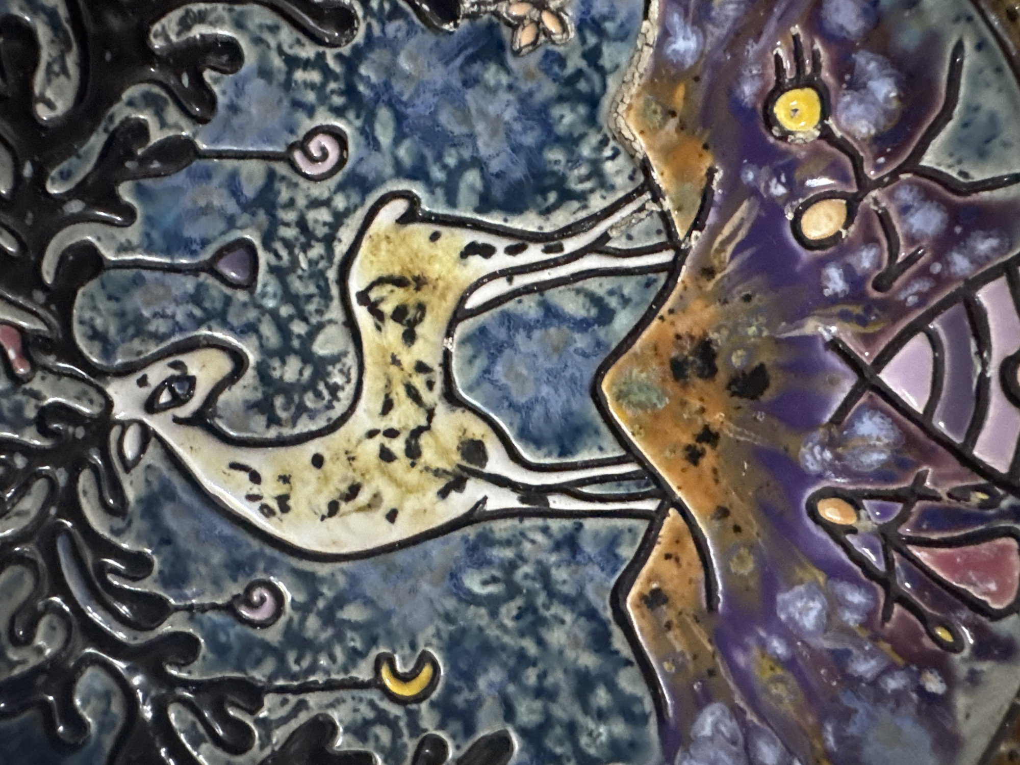 This is a close-up, detailed photograph of a ceramic tile with a whimsical, stylized depiction of a deer in a fantastical landscape. The deer is light-colored with dark spots and stands on a brown, textured hill. Behind the deer is a dark, gnarled tree with branches that suspend spiral shapes in shades of pink and purple, and a crescent moon in yellow. The background is a mottled blue and green. In the foreground, beneath the hill, is a landscape with purple, blue, and orange colors, featuring abstract figures and shapes. One prominent feature in the foreground is a creature or face with large, yellow eyes.
The image is a decorative ceramic tile, likely part of a larger artwork or functional item. It does not depict any specific location in Mũi Né, Vietnam.