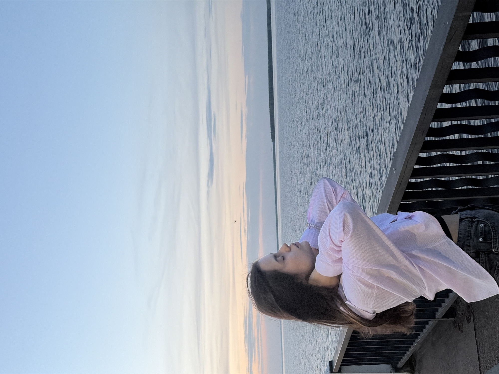 A woman lies on a dark metal staircase overlooking water. She wears a light pink, long-sleeved shirt and dark jeans. The background shows a sky gradient from blue to light orange and a body of water. The photograph was taken in the city of Chaikovsky, Russia.
