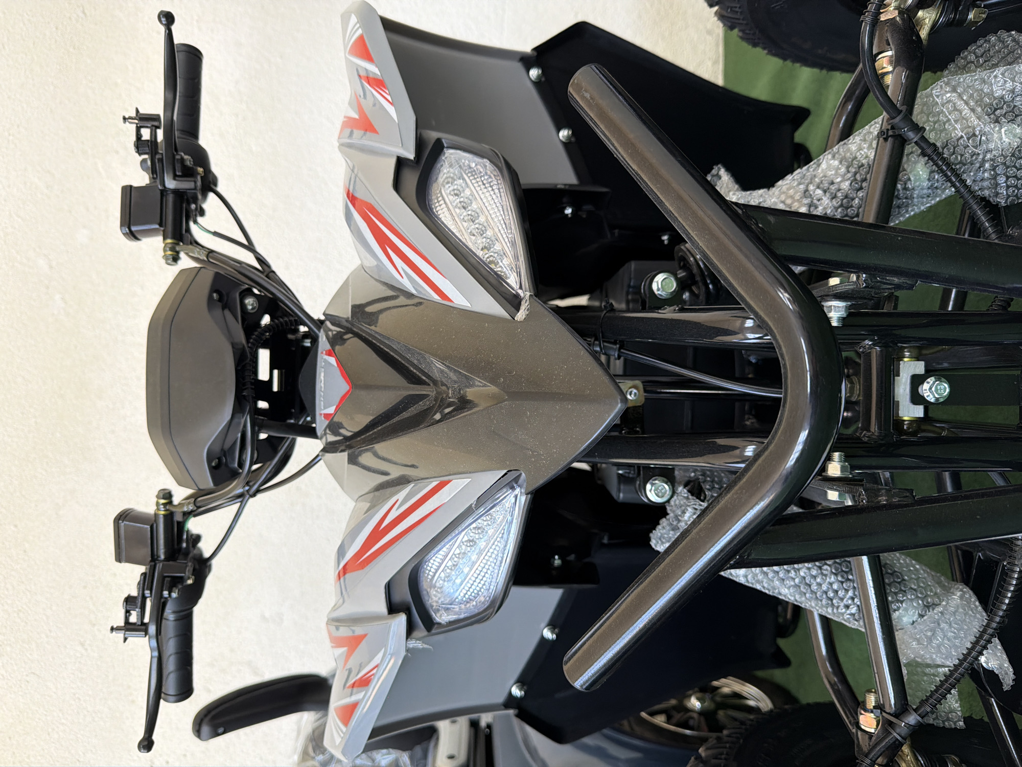 A front, eye-level shot of a grey and red ATV focuses on its handlebars and front fairing. The handlebars are black, with a brake lever and fluid reservoir visible on the right side. The ATV's front fairing is grey with red, angular decals. It features two clear, LED headlights. A black, curved metal bar is positioned in front of the ATV's lower section, obscuring some of the suspension components and wiring that are partially wrapped in bubble wrap. The ATV is positioned indoors against a light-colored wall, with a green surface visible on the ground.