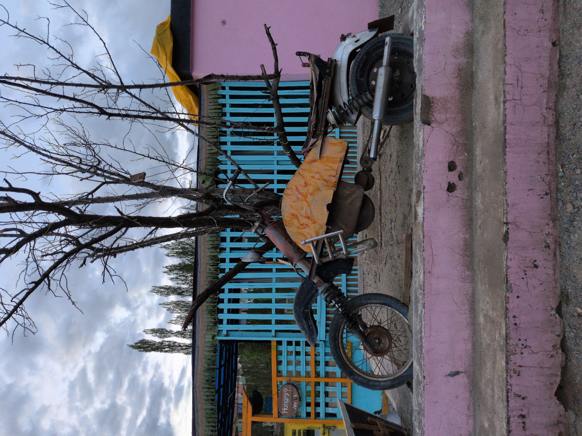 The image depicts a weathered, custom-built motorcycle displayed outdoors in Leh, India, near a pink-walled building and a turquoise wooden fence. The motorcycle, appearing to be an art installation rather than a functional vehicle, is made of various metal parts, including a rusted frame, a unique orange and yellow patterned fuel tank, and two distinct wheels. The front wheel is a spoked wheel with a flat tire, partially buried in the dirt. The rear wheel is a smaller, solid wheel with a chrome exhaust pipe. The motorcycle is positioned on a dirt ground in front of a concrete step painted pink.
Behind the motorcycle, a turquoise wooden fence with vertical planks separates the immediate area from the background. To the left of the fence, a yellow metal gate with a sign that reads "Hungry? TRY US" is visible. Beyond the fence, a row of tall trees with bare branches stands against a cloudy sky. The pink building in the background has a dark roof covered with a yellow tarp. The overall scene suggests a quirky, possibly commercial, outdoor display in a somewhat arid environment. There are no people visible in the image.