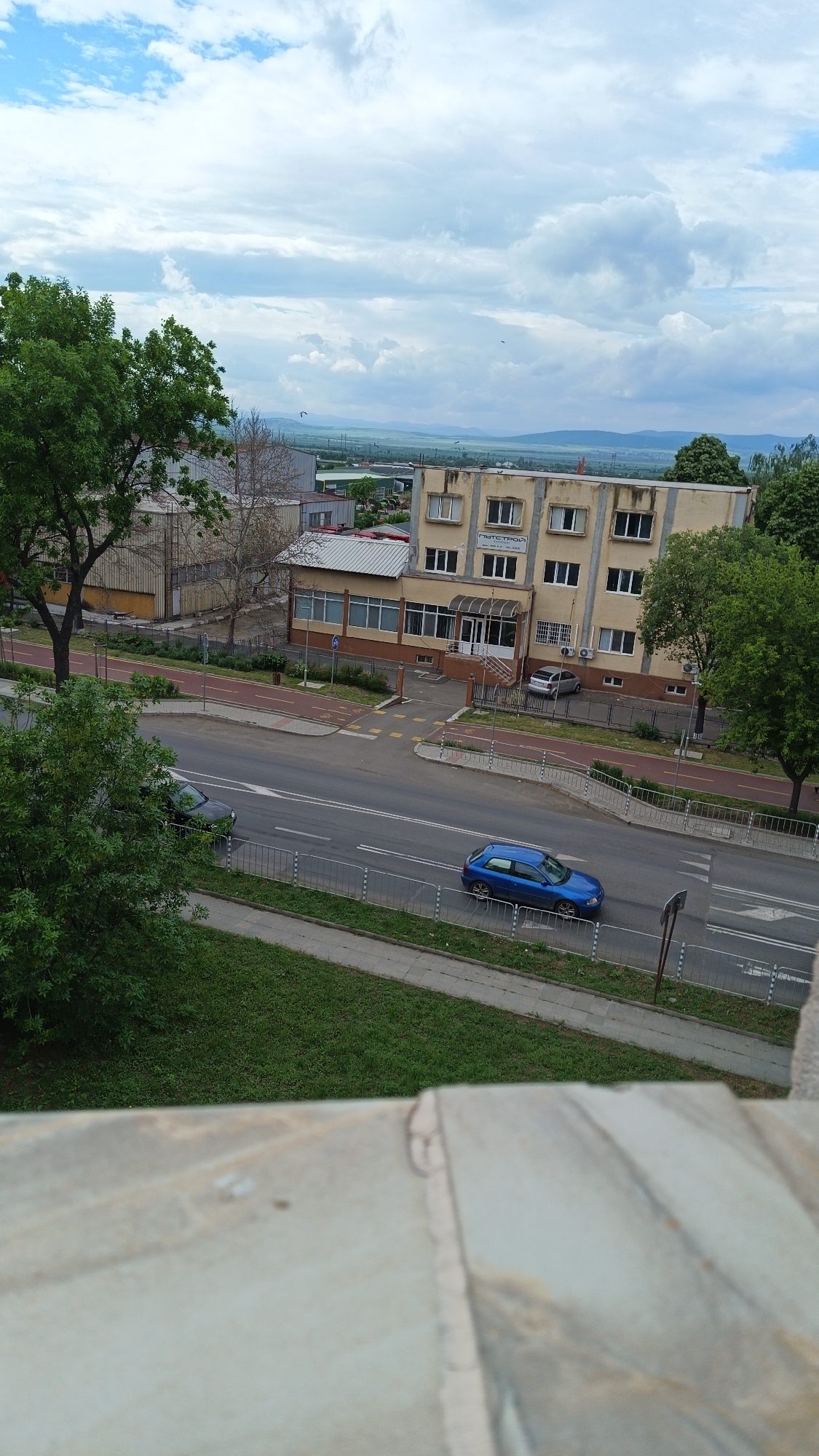 The image captures an elevated view of a street scene in Unknown, Unknown, under a sky with scattered clouds. The scene includes a road running horizontally across the frame, with two cars visible: a blue hatchback and a dark-colored sedan. Buildings border the road; one is a multi-story beige building with visible text on the facade, and another is a metal industrial building. Green trees and landscaping are present on either side of the road, with grass visible in the foreground and further background. A bike lane is situated adjacent to the road, marked in red. In the distance, there is a horizon with visible mountains or hills. A grey stone surface is visible at the very bottom of the image, likely indicating the location from which the photo was taken.