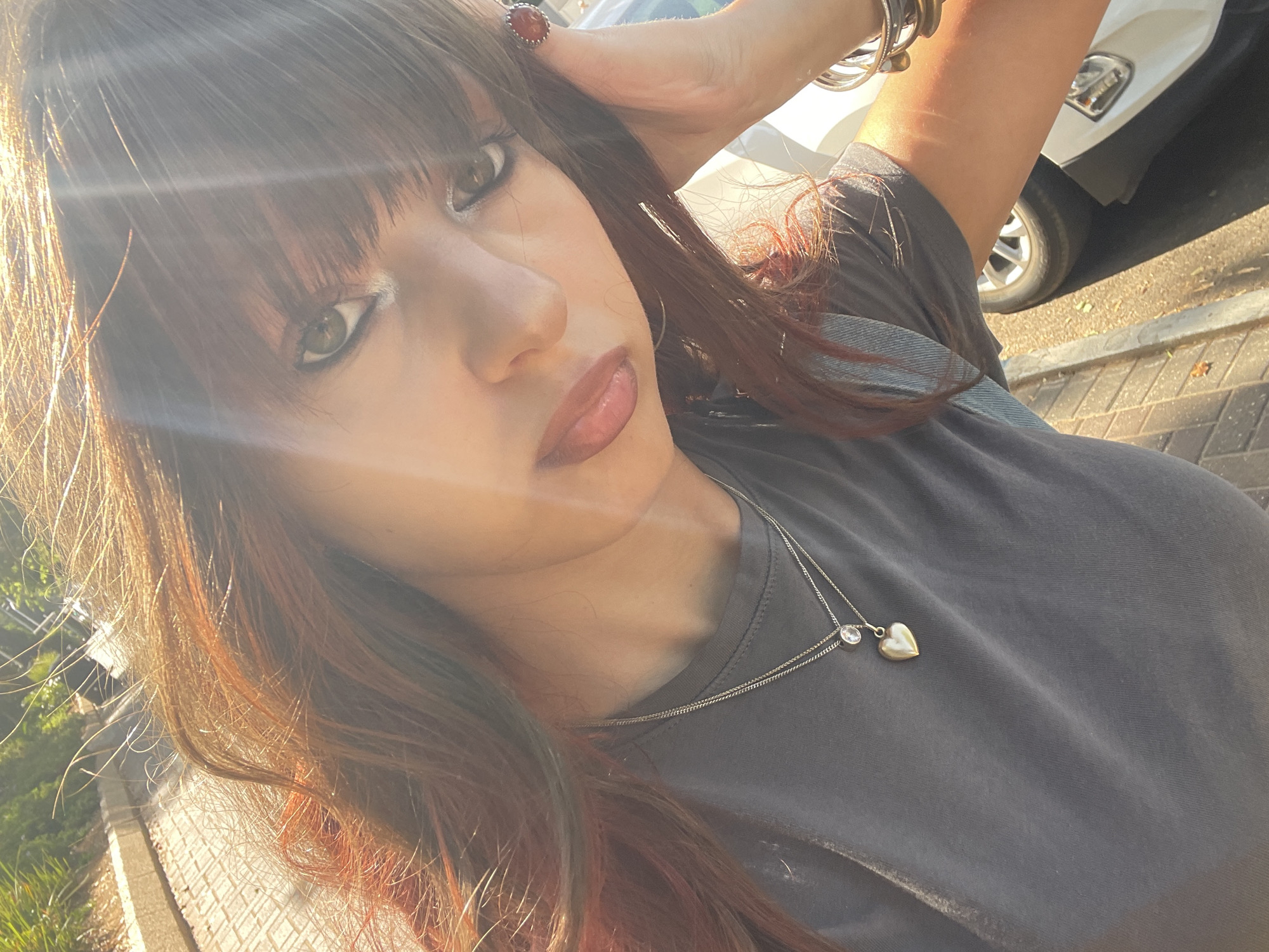 A close-up, selfie-style shot shows a young woman with long, reddish-brown hair, bangs, and green eyes. She is wearing a dark gray t-shirt with a necklace featuring a heart pendant and a small clear stone. She has on makeup, including eyeliner and lipstick. The woman is outdoors, with sunlight streaming from the upper right, creating lens flare and highlighting strands of her hair. Her right hand is raised, resting on her head and partially obscuring her ear, where she wears a ring with a reddish-orange stone. She has several bracelets on her left wrist. In the background, blurred elements suggest a street or sidewalk with brick paving and greenery. A portion of a white car is visible in the lower right. The scene is consistent with an outdoor setting in the city of Raanana, Israel.