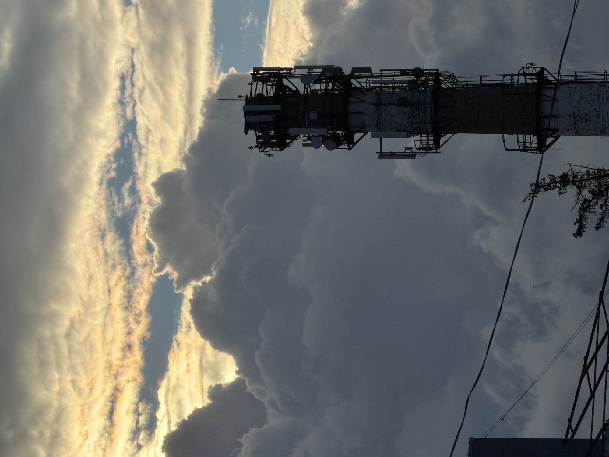 The image shows a tall telecommunications tower in Malakhovka, Russia. The tower is composed of a brick or concrete core structure with a metal framework extending outwards. Multiple antennas and equipment modules are mounted on the framework. The background is dominated by a sky filled with dense, billowing clouds, with some areas illuminated by sunlight. Electrical wires are visible in the lower right of the frame. Parts of a building are visible in the bottom of the image.