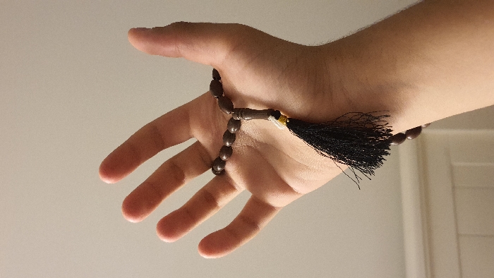 The image shows a person's right hand holding a brown wooden rosary with a black tassel, which is being held up in front of a white wall. The hand is partially visible, with the forearm and part of the upper arm also visible. The tassel of the rosary is hanging down from the hand, with some of the beads visible in the background. The location, based on the provided coordinates, is a residential area in Libourne, France.