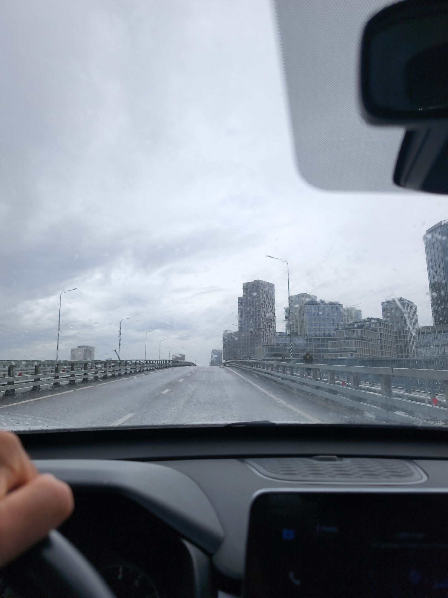 The image shows a view from inside a car driving on a bridge on a rainy day. The road is wet, and there are raindrops on the windshield. The bridge has metal railings on both sides. In the distance, there are several modern high-rise buildings, likely located in the city of Moscow, Russia. Streetlights line the bridge. Inside the car, part of the steering wheel and dashboard are visible. The rearview mirror is also in view. A hand is visible on the left side of the frame. The sky is overcast and gray.