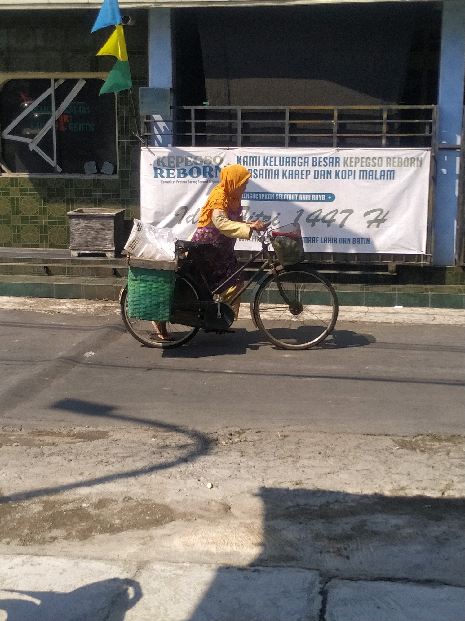 The image shows a woman wearing an orange hijab and a patterned dress riding a bicycle. She is carrying a green woven basket on the back of her bicycle, which contains various items, including what appears to be plastic containers. In front of the basket, a white plastic crate is also attached. She is riding on a paved road in front of a building with a banner that reads "KAMI KELUARGA BESAR KEPECSO REBORN" and "MENGUCAPKAN SELAMAT HARI RAYA". There are also flags flying above the building. The scene suggests a residential or community area in Batan, Indonesia, possibly during a festive occasion. The road surface shows signs of wear and tear, and shadows from a light source, likely the sun, are cast across the road and the foreground. The background of the building appears to have some greenery and a patterned wall.
