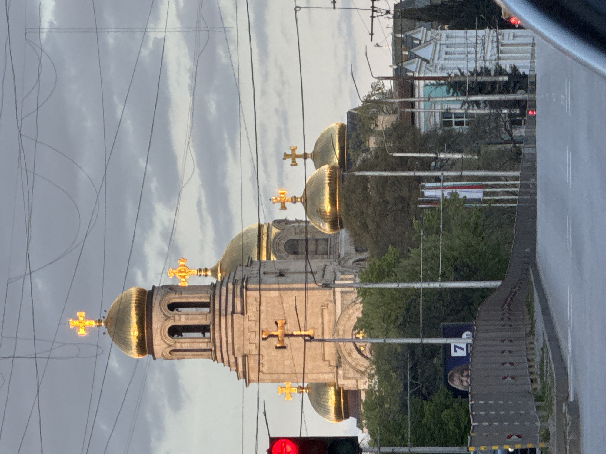 The image displays an urban scene in Varna, Bulgaria, dominated by a prominent Eastern Orthodox church featuring multiple golden domes and crosses. The church's main structure appears to be constructed from light-colored stone or stucco, with a tall bell tower topped by a golden dome and cross. Several other golden domes, also adorned with crosses, are visible behind and around the main tower. The domes brightly reflect light, contrasting with the overcast, cloudy sky.
Numerous overhead power lines and cables crisscross the upper and middle sections of the frame. Green trees and foliage are visible around the church and along the roadside. In the background, modern buildings are partially seen, one with intricate white structures that resemble scaffolding or a unique architectural facade.
In the foreground, a red traffic light is illuminated on the left. A paved road and an adjacent path or sidewalk stretch across the lower and right parts of the image. Further down the road, a billboard is visible, featuring a partial image of a person's face and the large white number "7", along with a partial "6" or "9", against a dark blue background. The lighting suggests it is either late afternoon or early morning on a day with significant cloud cover.