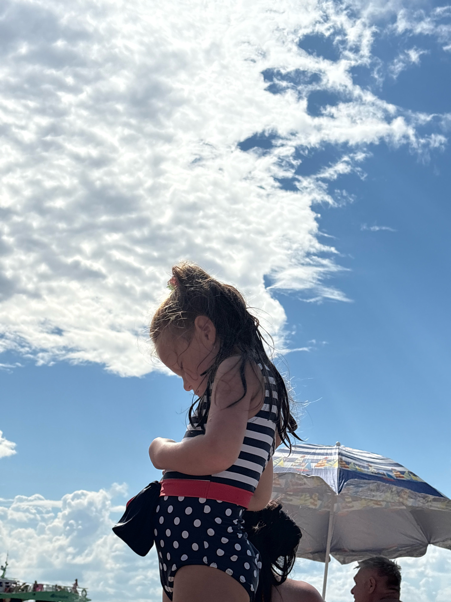 The image captures a child on a beach, likely in Arkhipo-Osipovka, Russia, under a bright, cloudy sky. The child, seen in profile, is wearing a striped and polka-dotted swimsuit. Their head is bowed, and they appear to be looking down at something in their hands. Their hair is wet, suggesting they may have been swimming or playing in the water.
Behind the child, an umbrella with a colorful nautical-themed pattern provides shade. A person's head and shoulders are visible in front of the umbrella, looking towards the right. Further in the background, a boat with people on board is partially visible on the left, suggesting a lively beach environment.
The sky is a prominent feature, filled with a mix of fluffy white clouds and patches of blue sky, indicating a partly cloudy day. The lighting suggests it is daytime, likely mid-morning or afternoon. The overall mood of the image is peaceful, focusing on the child's quiet moment amidst the activity of a beach day. There is no visible text in the image.