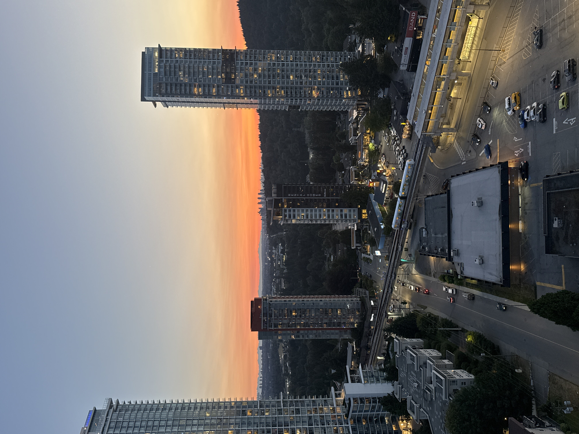 This is an aerial, high-angle, dusk photograph of a cityscape in Coquitlam, Canada. The sky displays a gradient from a pale blue at the top to a vibrant orange and yellow near the horizon, indicating sunset. Dominating the scene are several modern high-rise buildings, some with illuminated windows. In the foreground and mid-ground, a transit line with an elevated track and a train is visible. Several vehicles are present on roads and in parking lots, including cars and a truck. The environment includes dense green trees, commercial buildings with illuminated signage, and residential structures. The overall impression is a twilight view of an urban area transitioning into evening.