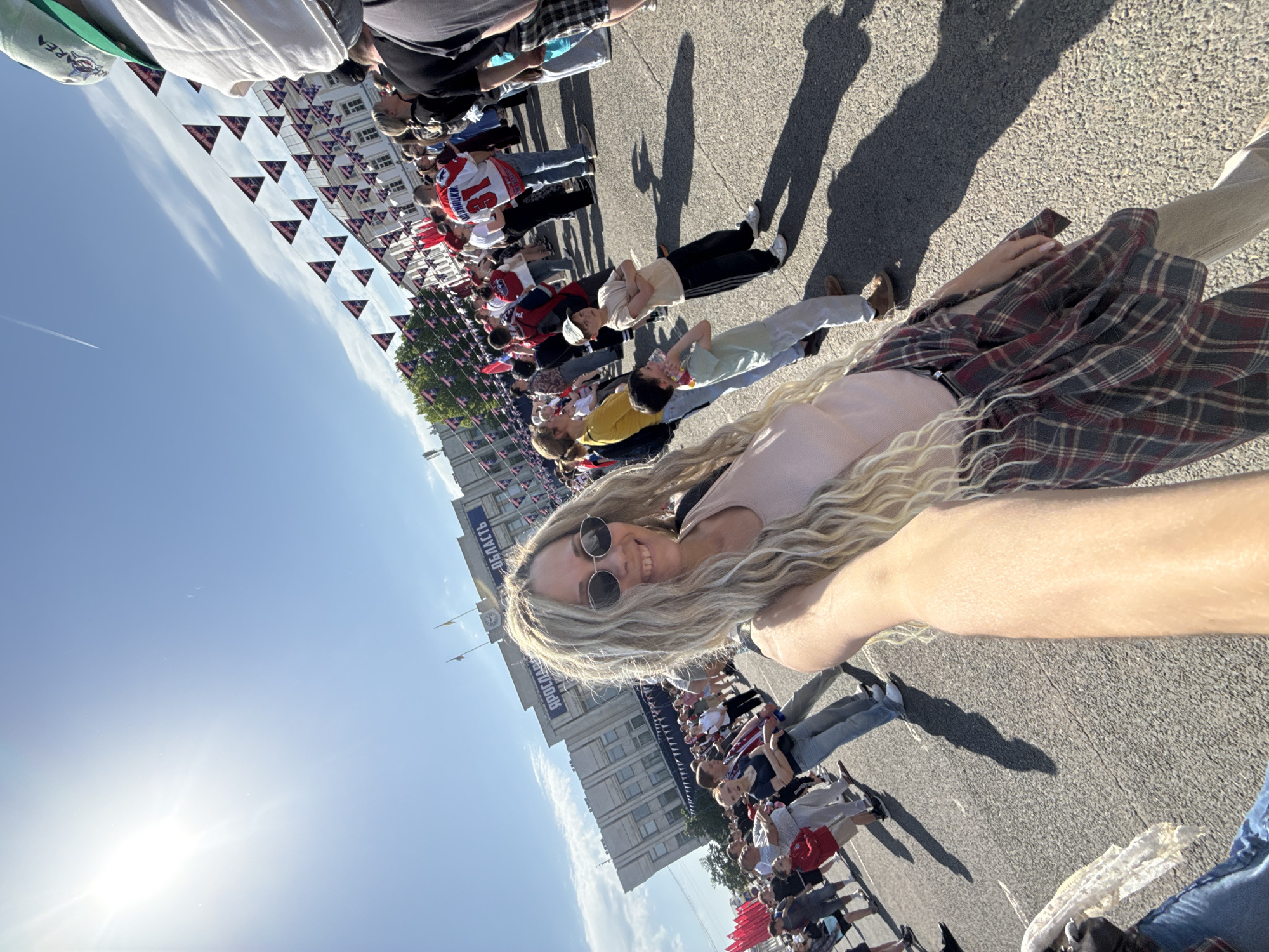 A medium shot captured from a low angle depicts a Caucasian woman smiling at the camera while standing in the street with people lining both sides and buildings in the background. The sky is clear and blue, with the sun shining brightly.
The woman is wearing round sunglasses, a pale pink top, and a brown and red plaid skirt, with a watch visible on her left wrist. She has long blonde wavy hair.
In the background, people are standing along the sides of the street, many of them dressed in casual attire. Some are wearing athletic jerseys and t-shirts. Many have flags.
A building with a Russian sign is visible in the backdrop. The street is lined with decorations of small red, white and blue triangular flags, adding a festive air to the event.
Shadows are cast on the street, suggesting it is a sunny day. The location appears to be a city in Russia, with a crowd gathered for what may be a parade or celebration.