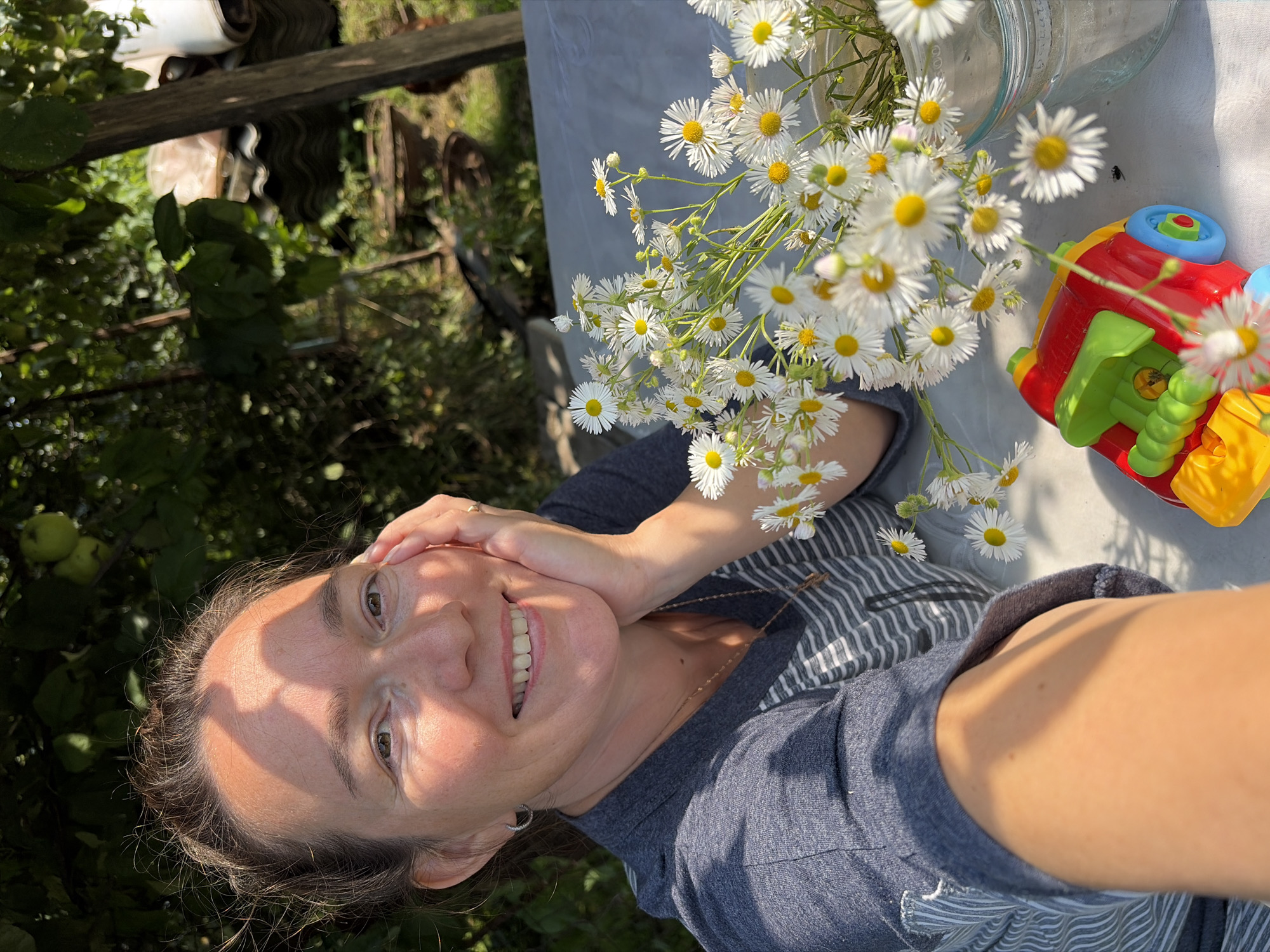 A selfie taken outdoors in Узяр, Russia. The image features a smiling woman in the foreground, with a bouquet of daisies and a toy train on a table. The background shows trees, a wooden post, and other elements of a yard. The woman is wearing a gray t-shirt with stripes. The lighting suggests it is daytime, with sunlight casting shadows.