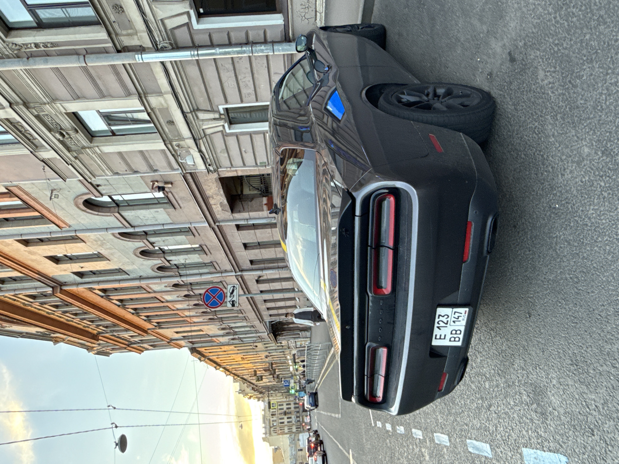 A black Dodge Challenger is parked on a city street in Saint Petersburg, Russia. The car is viewed from the rear, with its license plate visible. The license plate reads "E 123 BB 147 RUS". The car has a yellow stripe down the middle of its trunk and a blue stripe on its rear fender. In the background, there are ornate buildings with arched windows and balconies, typical of St. Petersburg architecture. A traffic sign indicating no stopping is visible on the side of the road. The sky is partly cloudy and appears to be late afternoon or early evening, with warm light illuminating the buildings. Other cars are visible in the distance, and there are streetlights and electrical wires overhead. A pedestrian is walking on the sidewalk in front of the buildings. The street is paved and has lane markings.