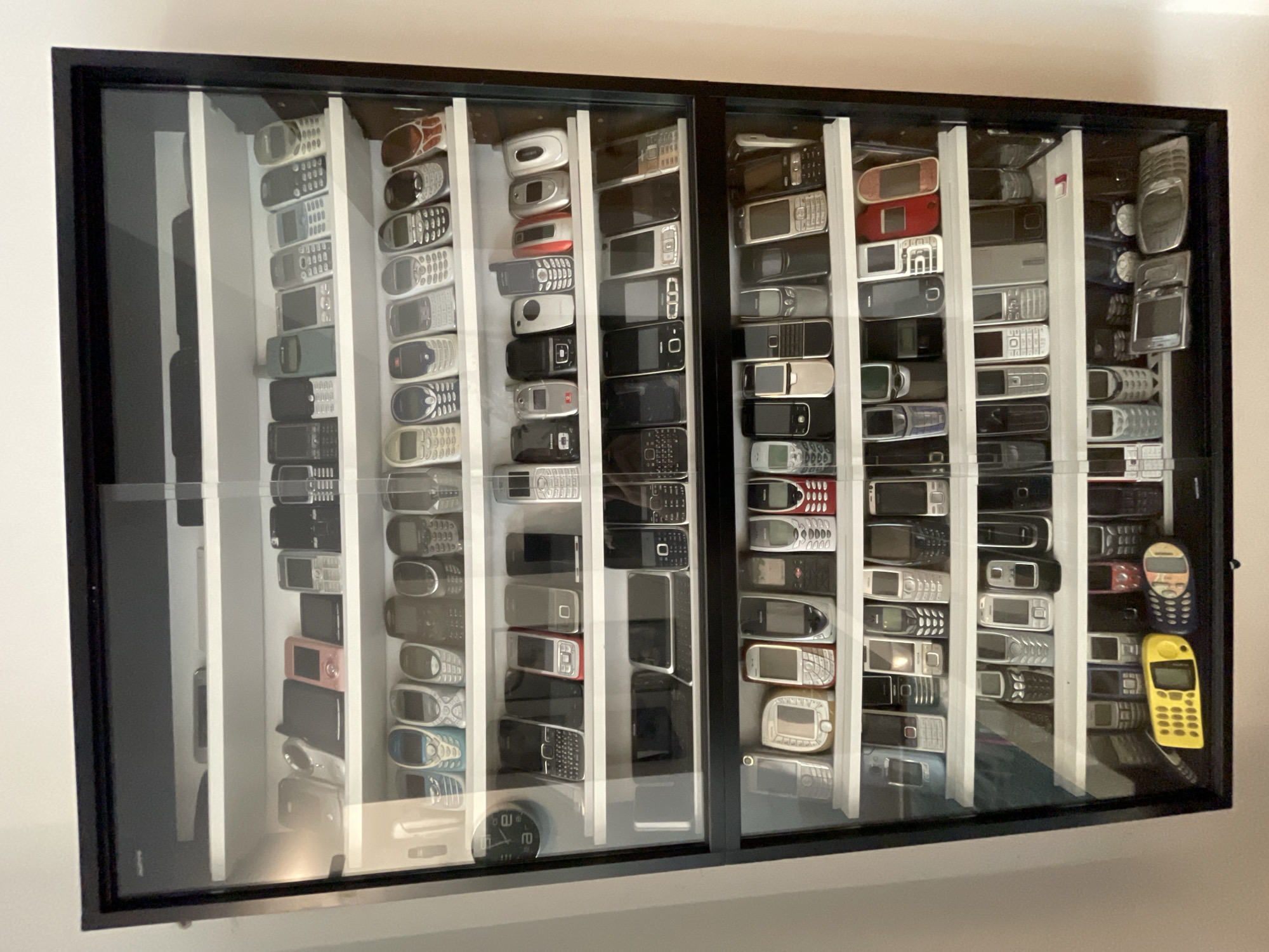 The image shows a display cabinet containing a large collection of mobile phones, primarily older models. The cabinet has a black frame and sliding glass doors. It is divided into multiple shelves, each filled with rows of phones. The phones vary in color, size, and design, including flip phones, candy bar phones, and some with slide-out keypads. The background is a plain white wall. Based on image analysis, this scene is located in Csömör, Hungary, specifically in a residential setting. There are no people visible in the image, and no actions or interactions are depicted. The environment is clean and organized, suggesting a display or collection rather than active use.