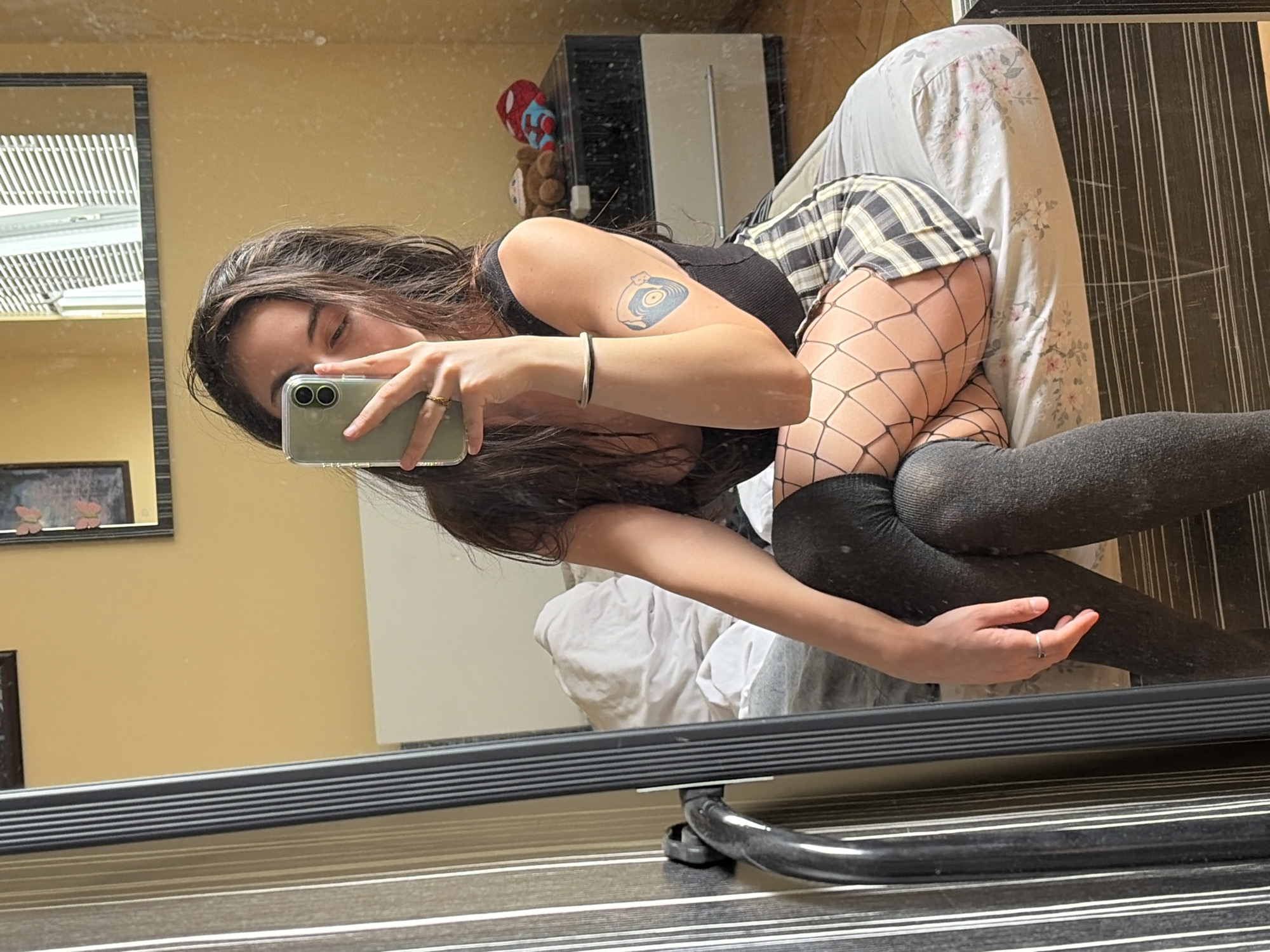 A young woman with long, dark hair is taking a selfie in a bedroom. She is wearing a black sleeveless top, a plaid skirt, and dark stockings with a fishnet pattern. She is also wearing a ring on her left hand and a bracelet on her right wrist. A tattoo of a record is visible on her right arm. The room has yellow walls and a bed with a floral patterned comforter. A mirror reflects the room, showing a window with blinds and a dark dresser with stuffed animals on top. The woman is sitting on the edge of the bed, holding a green smartphone in her right hand to take the photo.