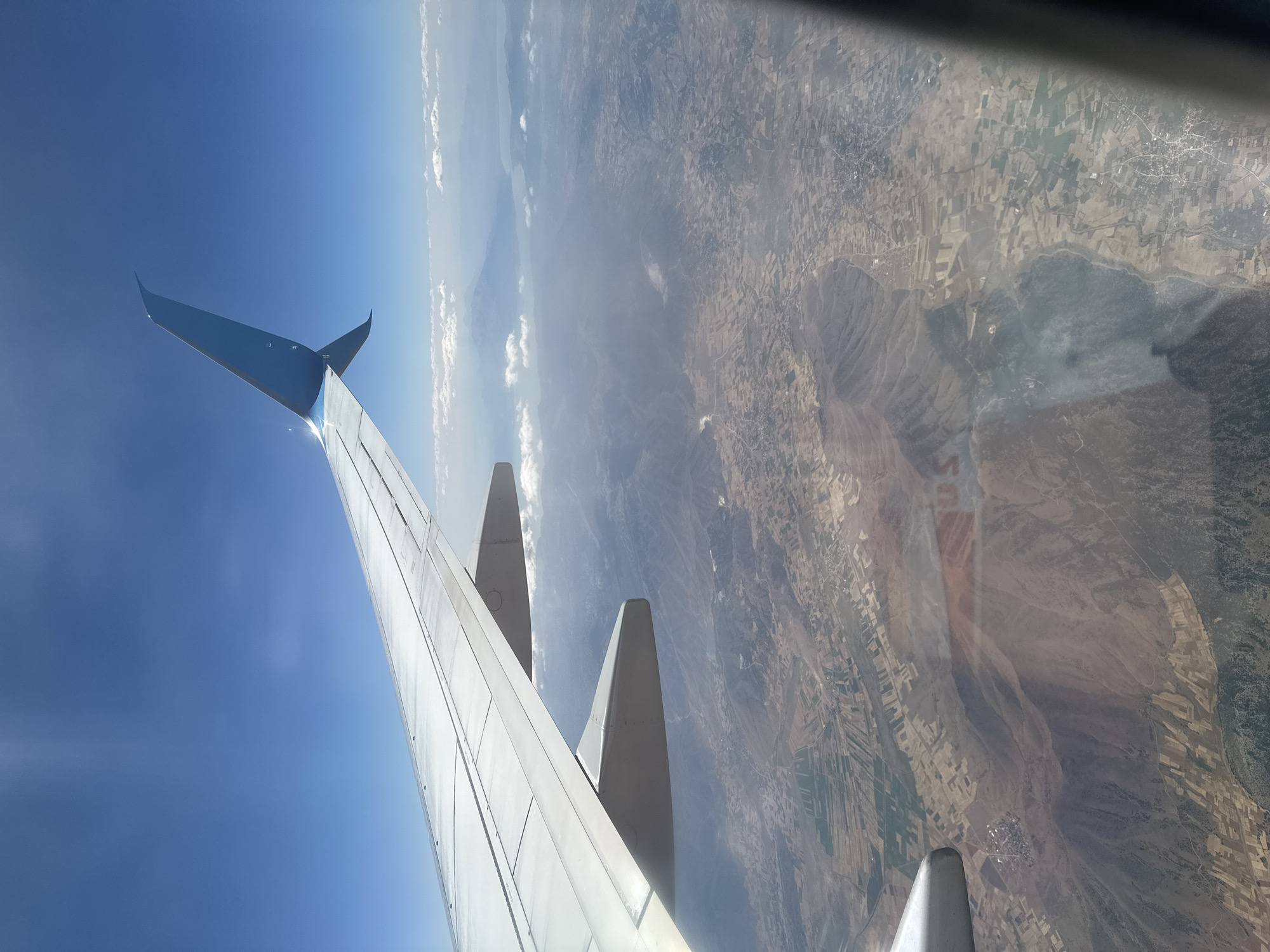 This is an aerial photograph taken from an airplane window, looking down at a landscape. The dominant feature in the foreground is the wing of the aircraft, painted white with a blue wingtip. The wing extends from the left side of the image across the upper portion. Below the wing, a vast, arid landscape unfolds, characterized by dry, brown earth, interspersed with fields of varying shades of green and tan, suggesting agricultural activity. Rolling hills and mountains are visible in the distance, some shrouded in haze. In the far distance, a body of water, likely a lake or sea, is visible, with a distant landmass or mountain range on the horizon. The sky is a clear, bright blue with a few wispy white clouds. The image appears to be taken during daylight hours, with the sun casting light and shadows on the landscape. The specific location in Şarkikaraağaç, Turkey, cannot be definitively identified from the visual information alone without external data.