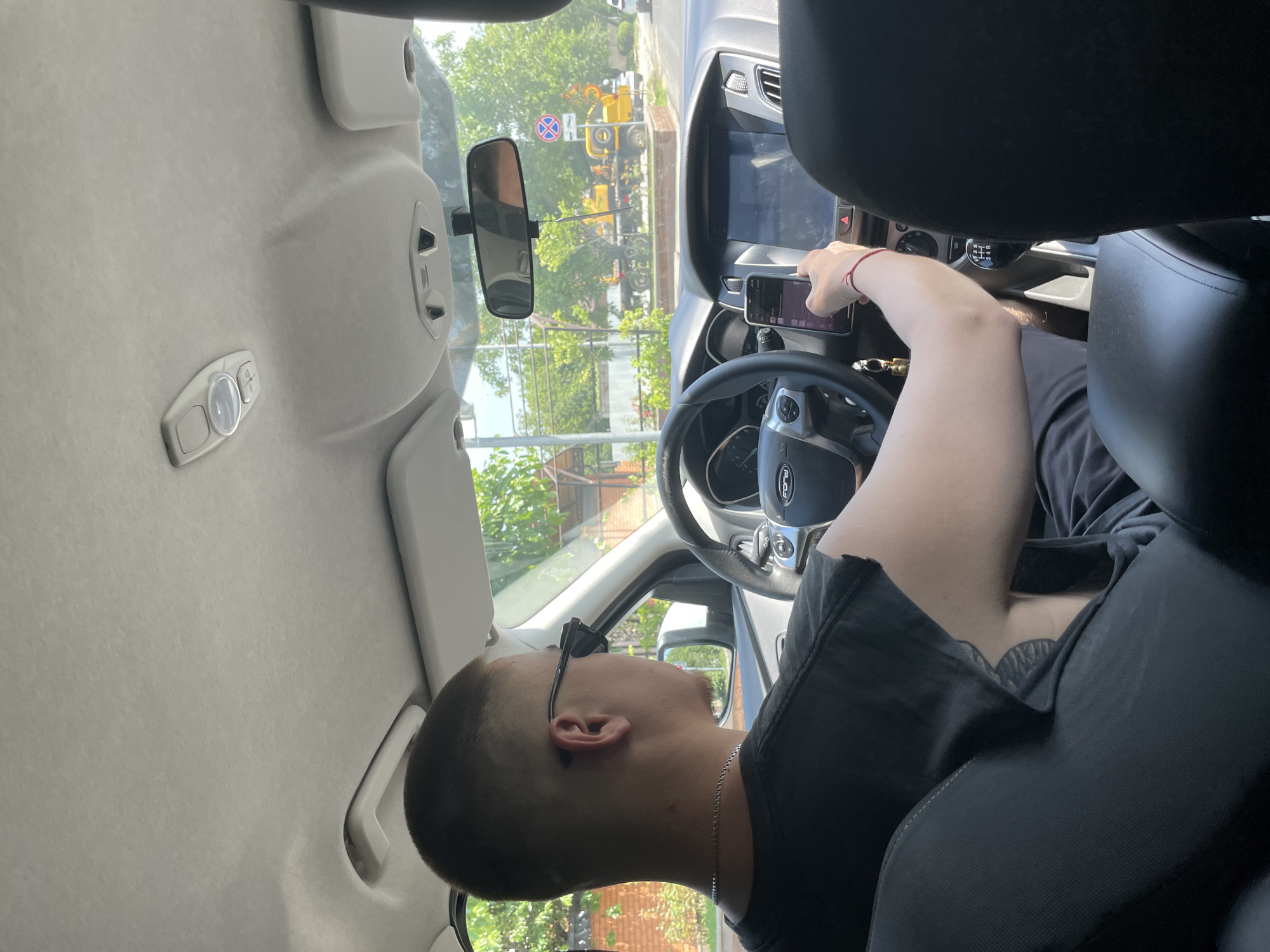 A man with a short haircut and sunglasses is driving a car. He is wearing a black t-shirt and a silver necklace. He is holding a smartphone in his right hand and appears to be looking at it. The car's dashboard is visible, including the steering wheel, infotainment screen, and gear shift. Outside the car, trees and buildings can be seen, suggesting an urban or suburban environment. The image is taken from the back seat of the car, looking forward.