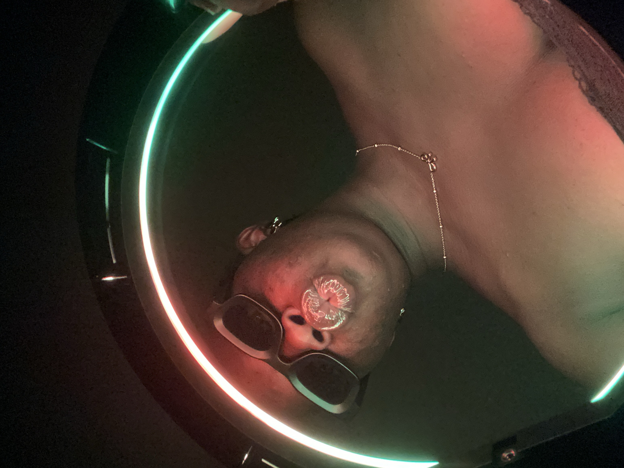 An inverted, medium close-up shot features a person's head and upper torso, illuminated by a dynamic neon light source. The individual has dark skin and is wearing dark sunglasses with thick, light-colored frames. A delicate necklace with a small, stylized pendant is visible around their neck. A highly reflective, clear, and textured substance covers their puckered lips, creating a striking, almost sculptural effect. Earrings are also visible.
The subject is framed by a prominent, curved LED light strip that transitions in color from a vibrant red-orange on the lower left to a cool green-aqua on the upper right. This colorful illumination casts strong hues onto the person's skin, particularly on the face and neck. The background is largely dark and unidentifiable, suggesting an indoor setting with controlled lighting. The person appears to be posing for a photograph, possibly a selfie, given the close proximity and deliberate styling, including the distinctive lip treatment and sunglasses. The overall mood is modern and artistic. There is no visible text. This scene takes place in Maigizo, Nigeria.