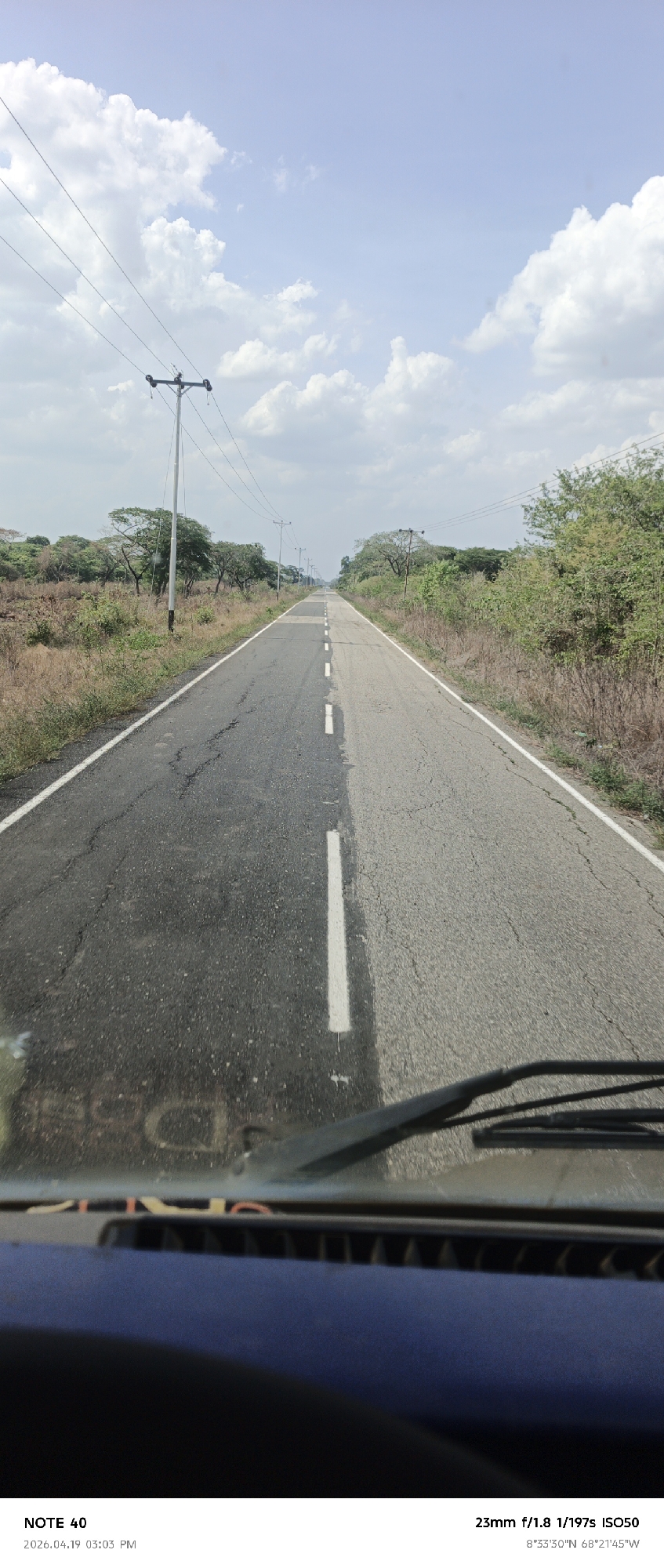 Here's a description of the image:
The image shows a straight asphalt road stretching into the distance under a partly cloudy sky. The road is divided by a white center line and flanked by vegetation on either side. There are telephone poles lining the road. The photograph appears to have been taken from inside a vehicle, with the dashboard and windshield wipers visible at the bottom of the frame. The image metadata indicates the location is 8°33'30"N, 68°21'45"W. Based on the metadata, the location is near Arismendi, Venezuela.